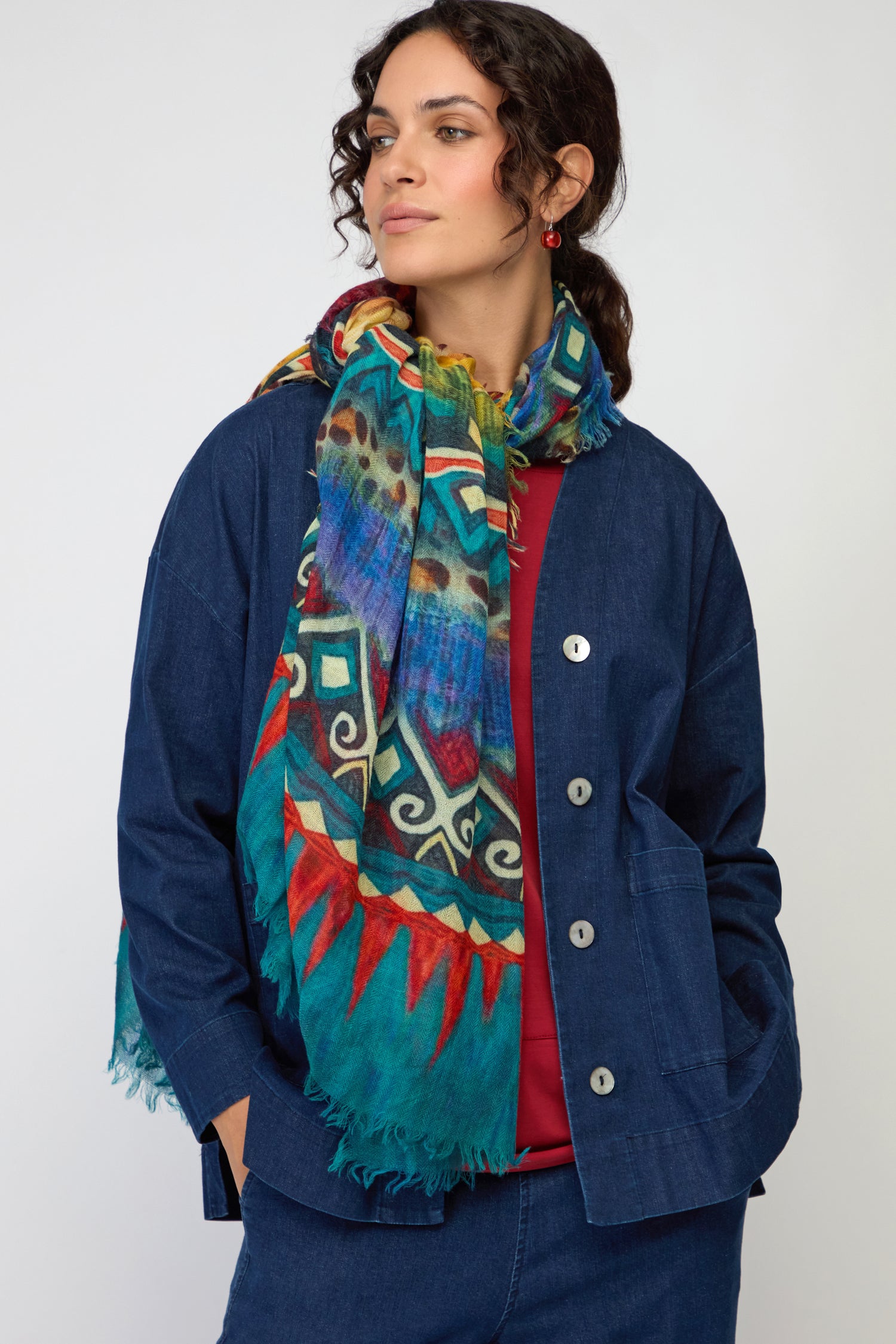A woman stands against a plain background wearing a blue jacket, jeans, and the Savannah Print Wool Scarf styled as a colorful patterned wrap around her neck.