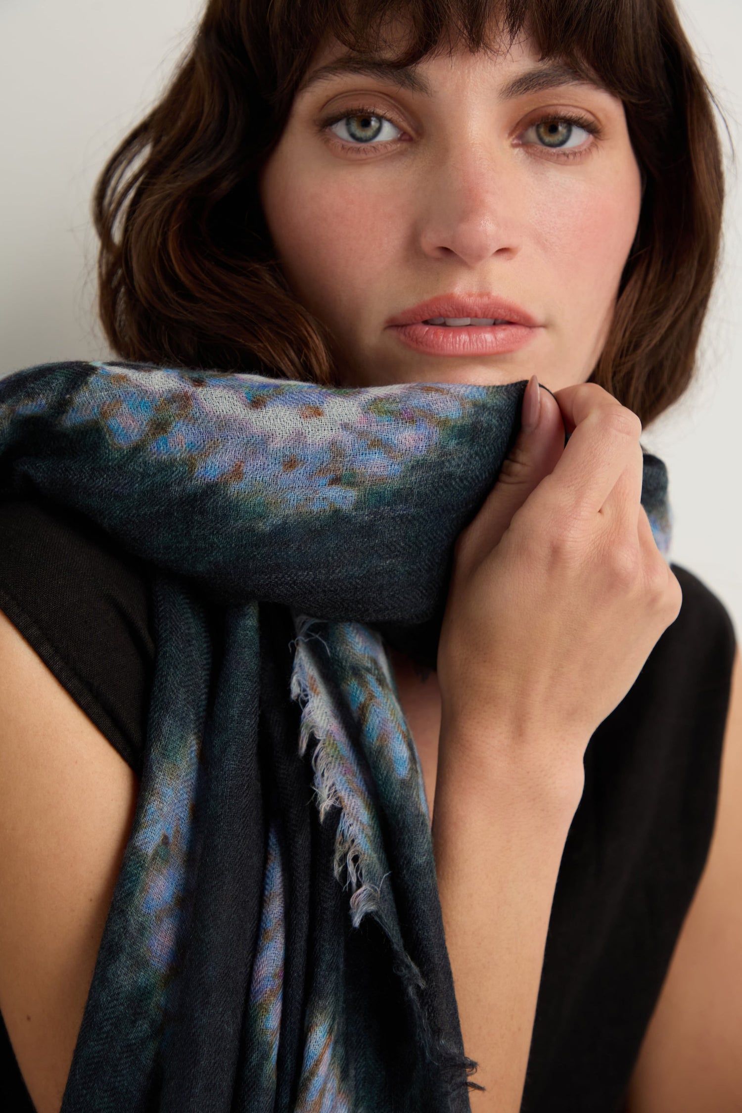 A woman with wavy brown hair holds the Shibori Wool Scarf close to her face. Wearing a black top, she gazes directly at the camera against a plain background, highlighting the breathable elegance of this accessory.