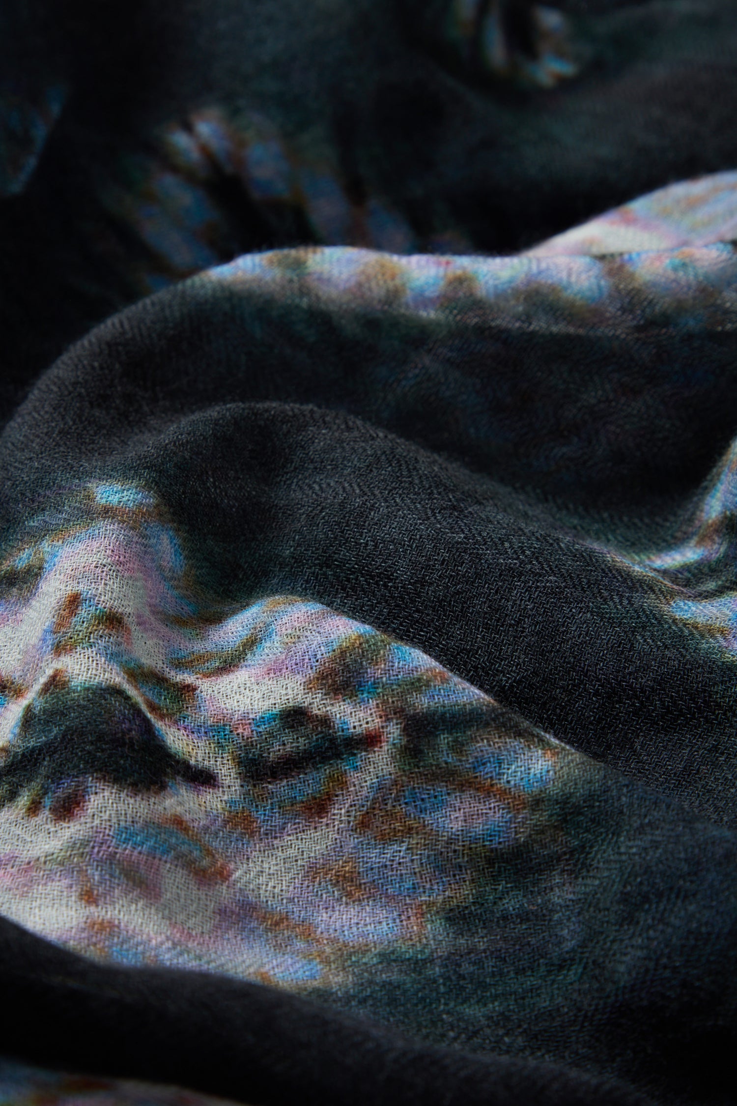 A close-up of the Shibori Wool Scarf reveals a textured fabric with a multicolored abstract pattern, featuring dark, wavy folds and subtle hints of blue, pink, and white. This scarf showcases a breathable design that blends artisanal style with comfort.