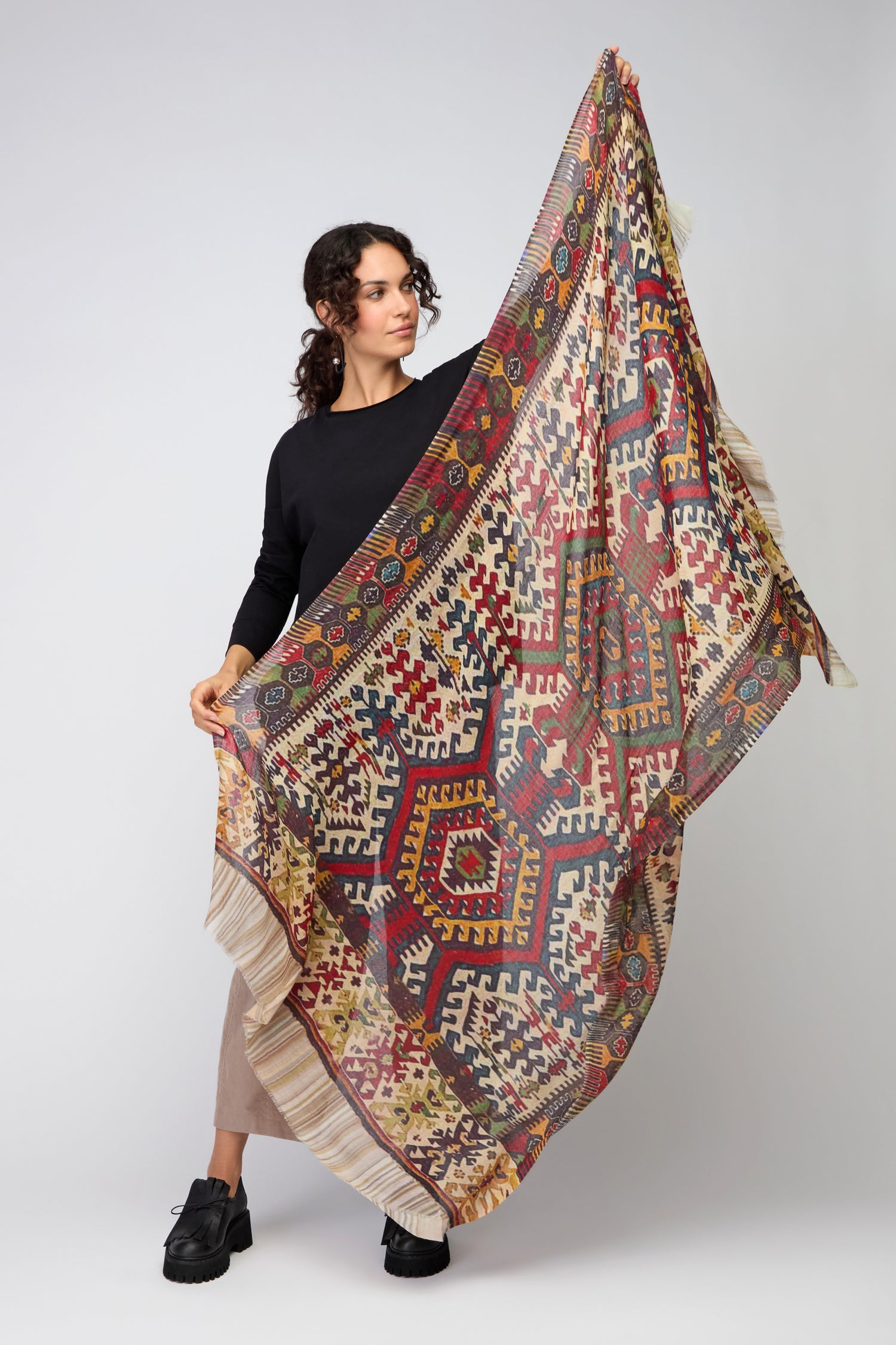 A woman displays the Tapestry Wool Scarf, featuring large geometric patterns in red, yellow, and black tones, against a plain light background.