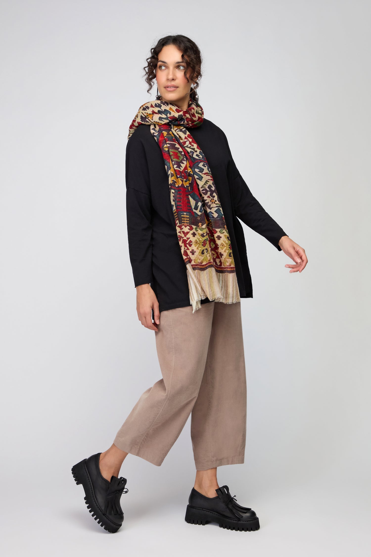 A woman stands against a plain light background, wearing a black long-sleeve top, beige wide-leg pants, black platform shoes, and the Tapestry Wool Scarf.