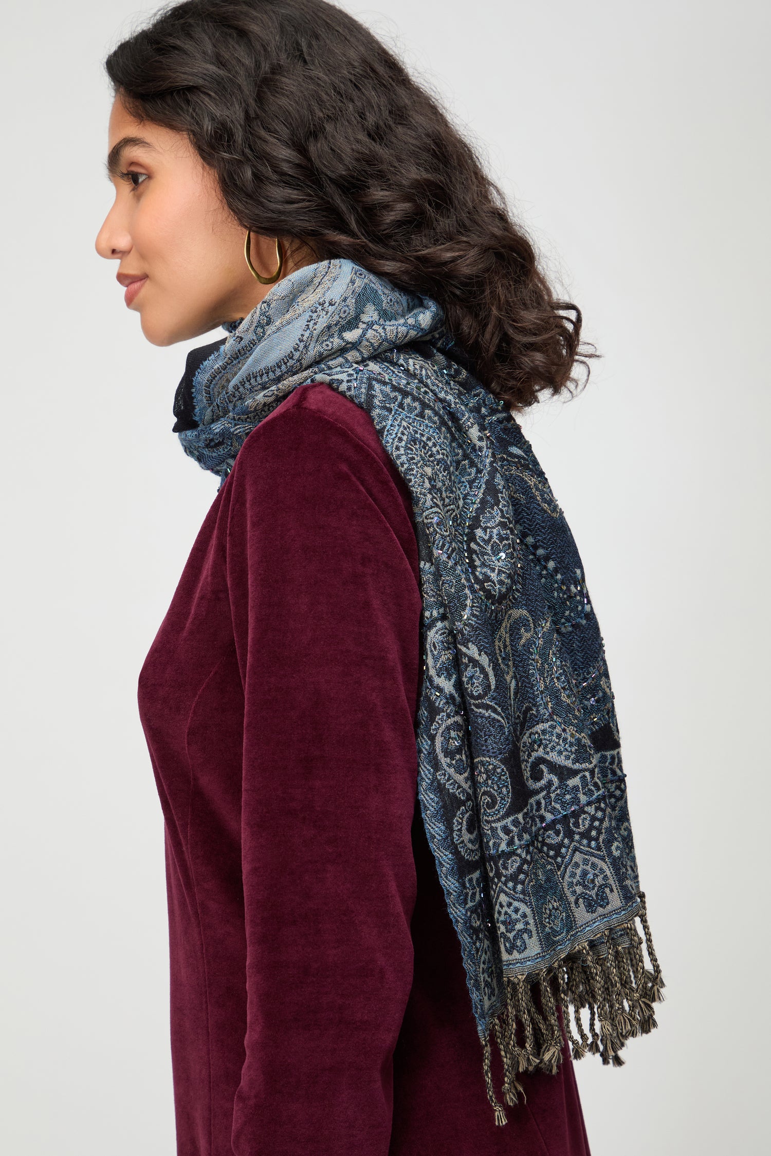 A woman in a burgundy velvet top and gold hoop earrings wears the Opulent Paisley Wool Scarf with fringe, shown in profile against a plain background.