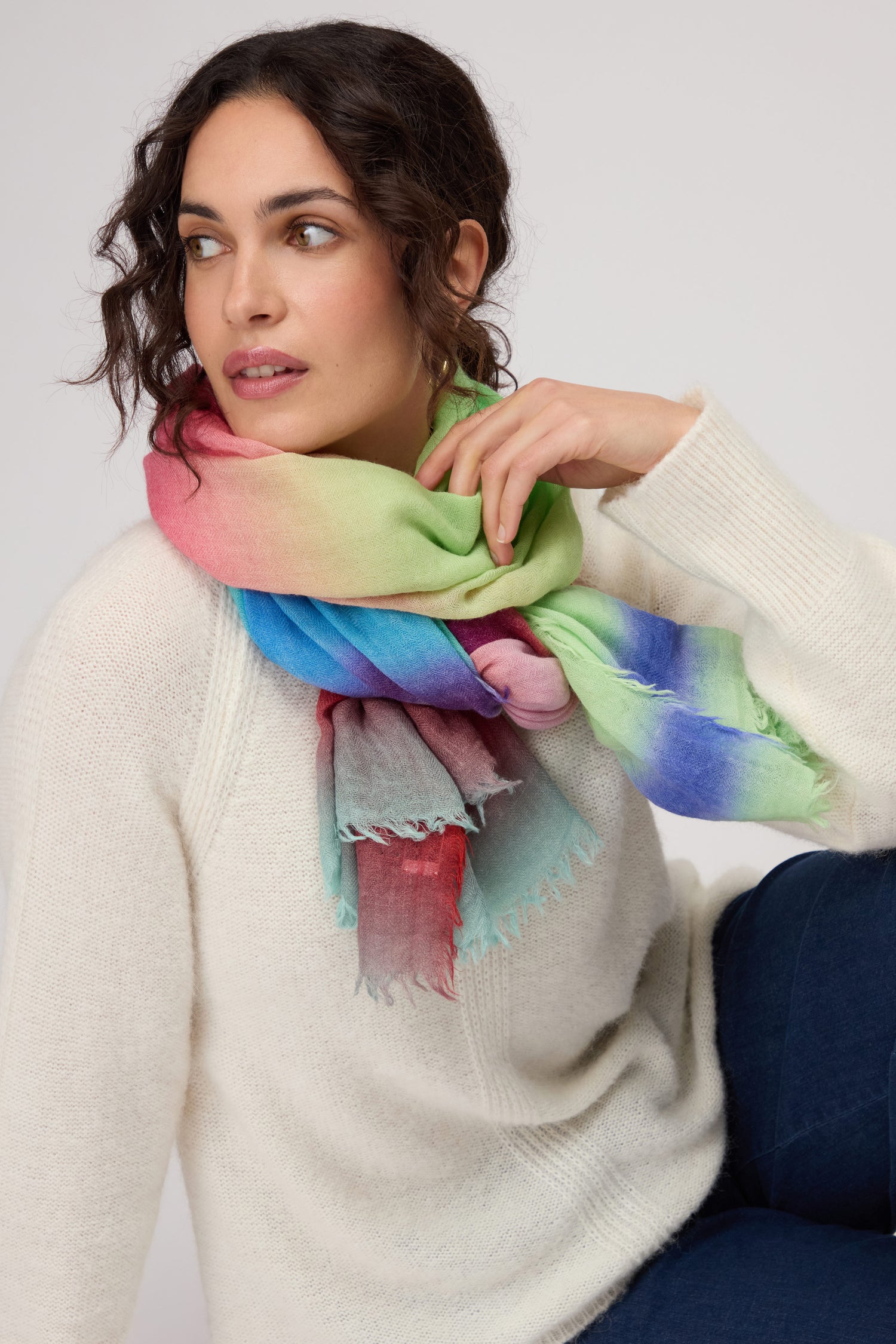 A woman in a cream sweater and dark blue jeans wears the Horizon Stripe Wool Scarf, featuring a lightweight ombre design, elegantly wrapped around her neck as she gazes to the side.