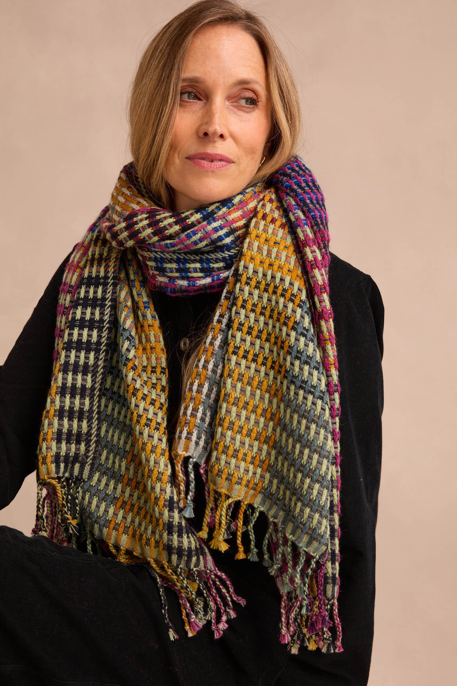 A woman wears the Checkerboard Scarf—an oversized, multicolored accessory—draped over a black coat. Seated with her arms crossed and gazing slightly to the side against a neutral background, she lets the lightweight wool-blend scarf add warmth and a vibrant pop to her ensemble.