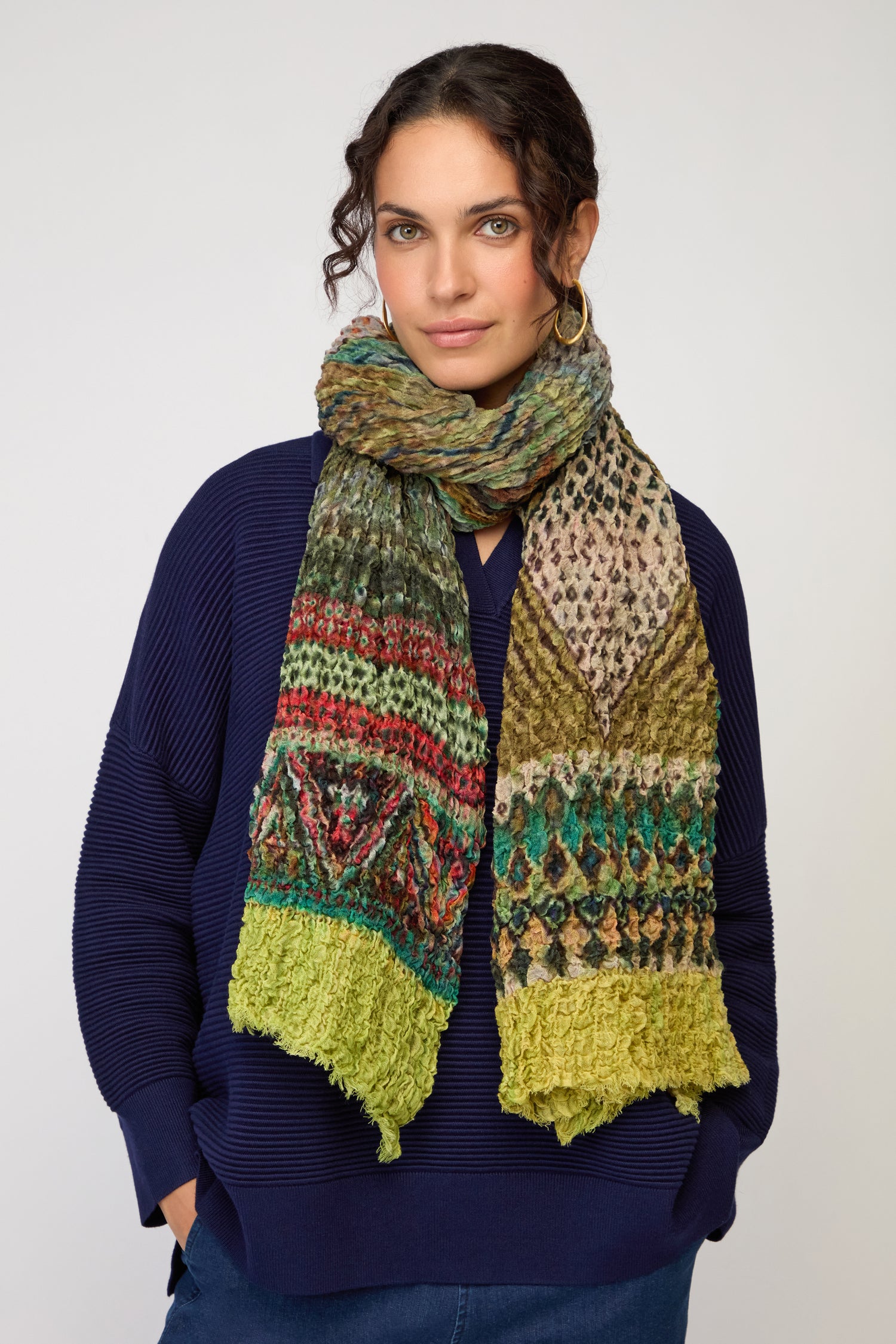 A woman stands against a plain light background, wearing a blue sweater and the Crushed Digital Print Wool Scarf, which features a multicolored pattern with green ends.
