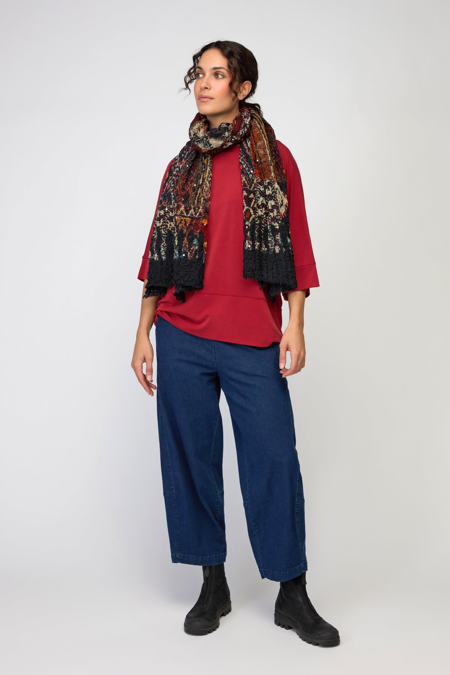 A person stands against a white background, wearing a red top, blue wide-leg jeans, black boots, and the Crushed Patchwork Sequin Scarf.