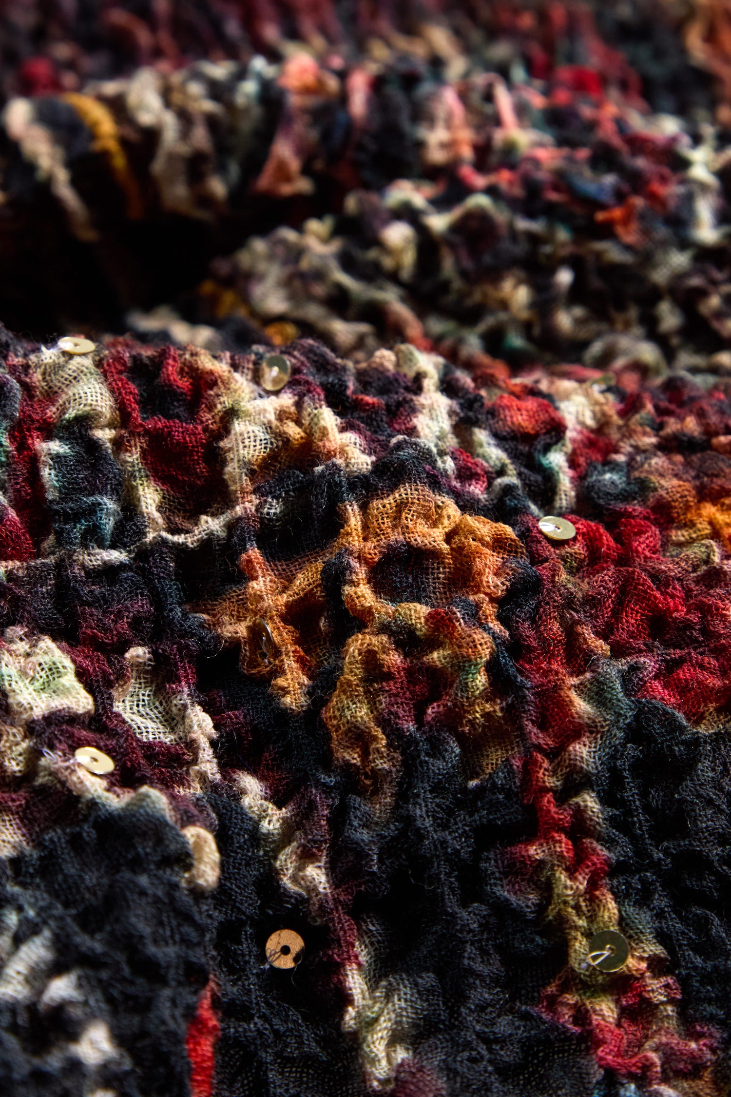 Close-up of the Crushed Patchwork Sequin Scarf, featuring textured, multicolored fabric with dark and warm tones and small round sequins—ideal for an oversized, wool-rich accessory.