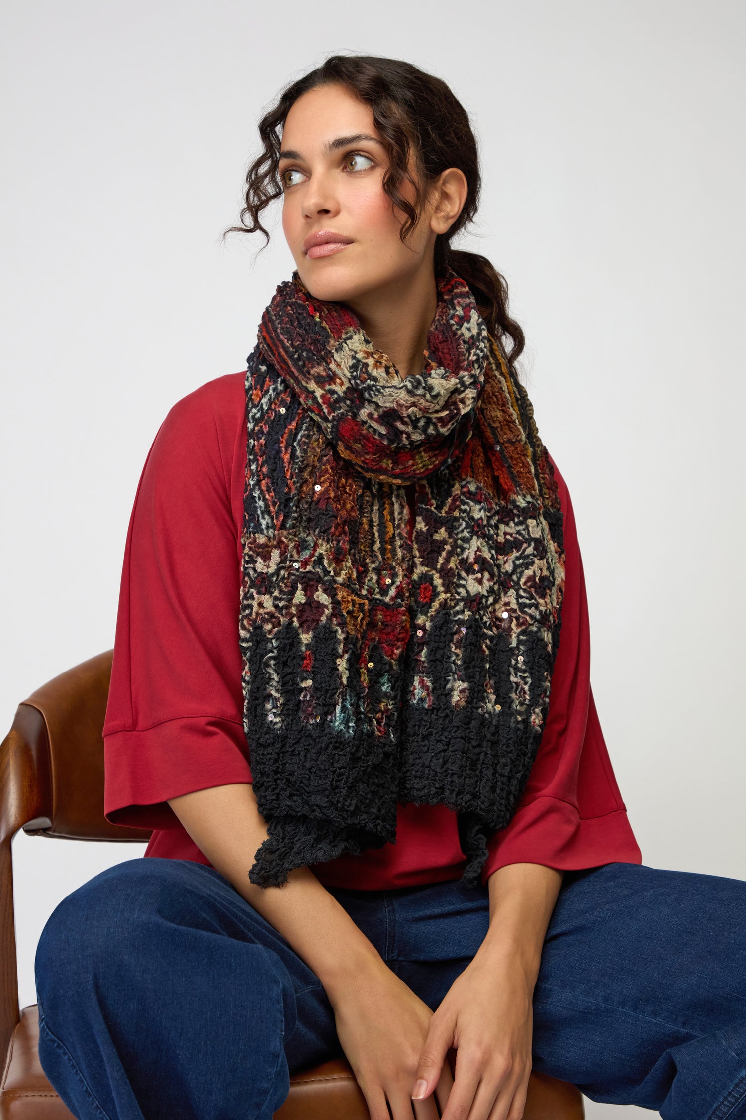 A woman sits on a brown chair wearing a red top, blue pants, and the Crushed Patchwork Sequin Scarf, looking to the side against a plain background.