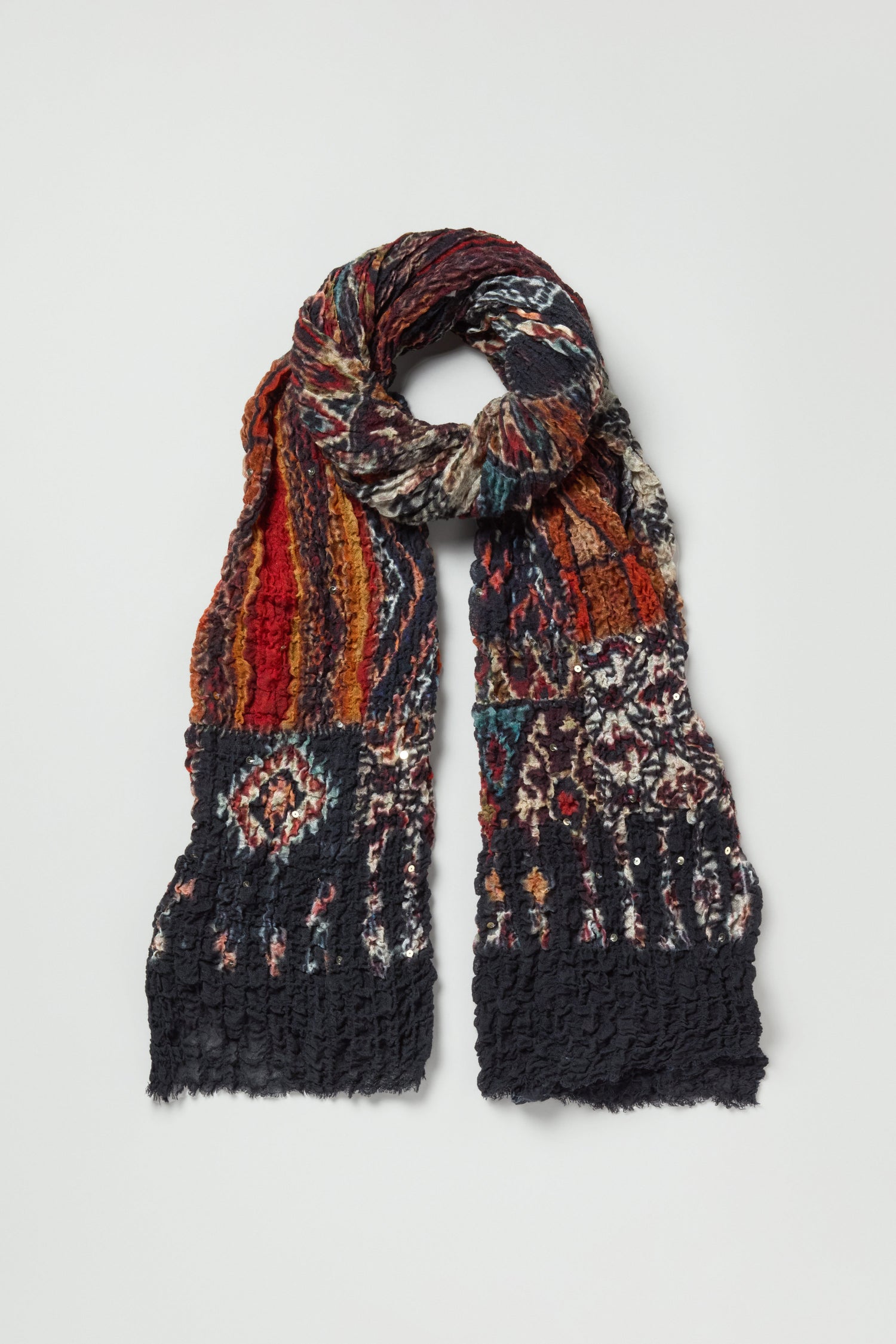 The Crushed Patchwork Sequin Scarf features multicolored geometric patterns in red, orange, blue, and black tones with a wool-rich texture and slightly fringed edge.