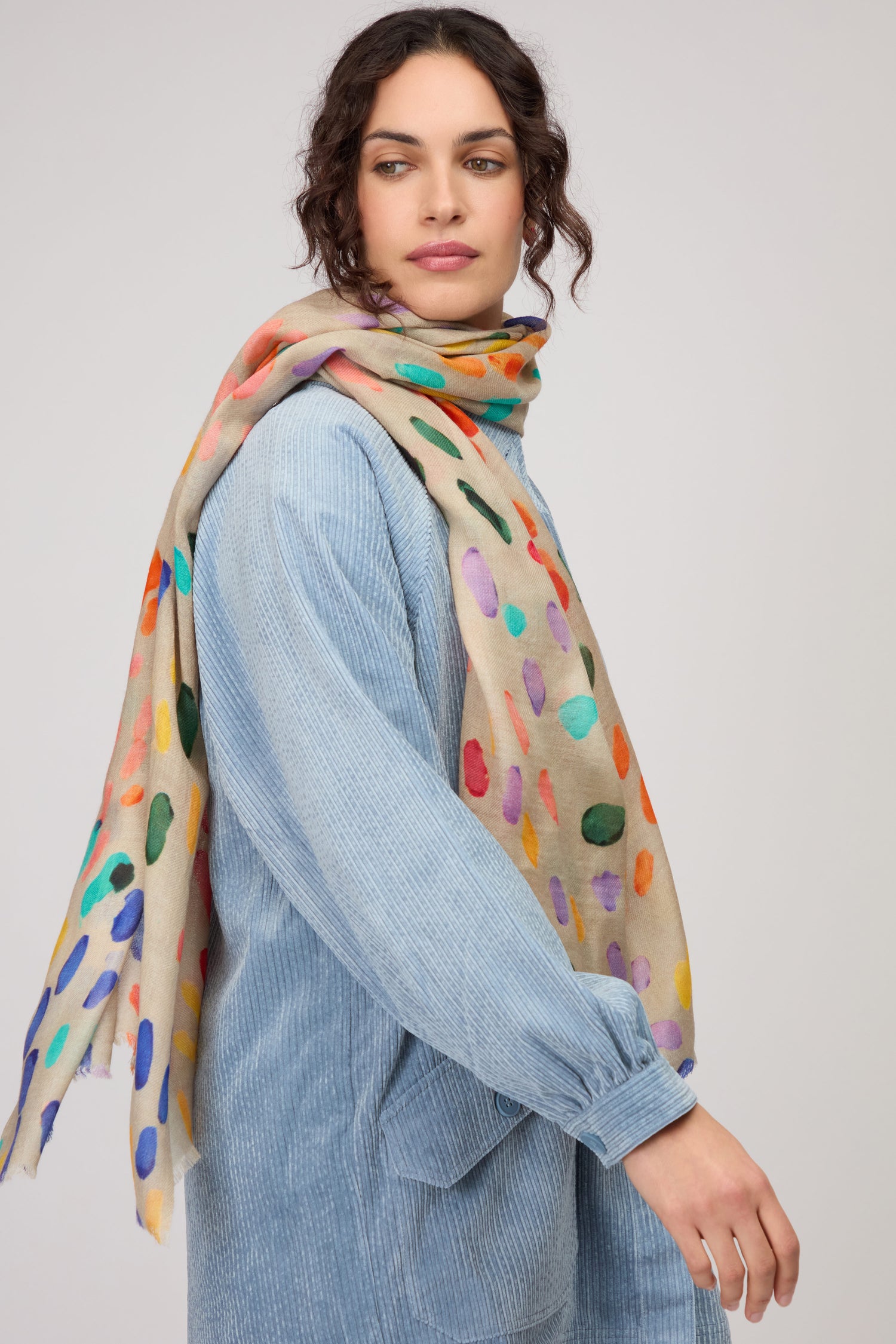 A woman stands against a plain light background wearing a light blue textured dress and the Artful Dots Wool Silk Scarf, featuring a colorful pattern.