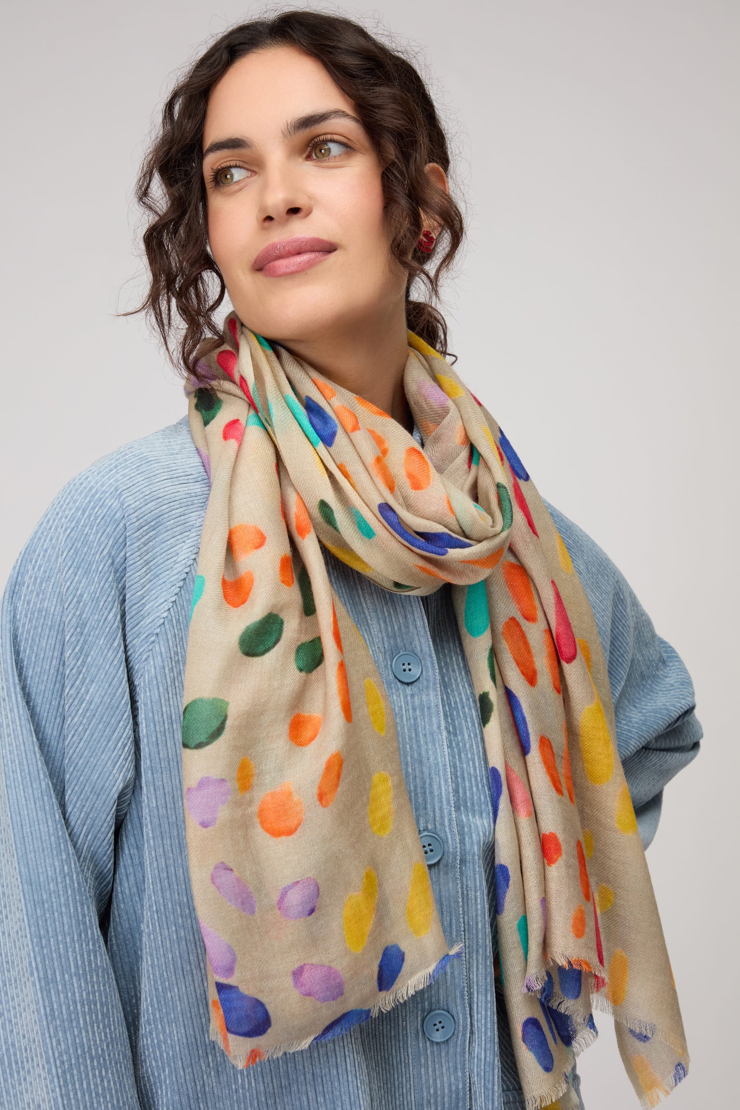 A woman wears the Artful Dots Wool Silk Scarf, featuring a colorful polka dot pattern, with a light blue textured button-up top, looking to the side against a neutral background.