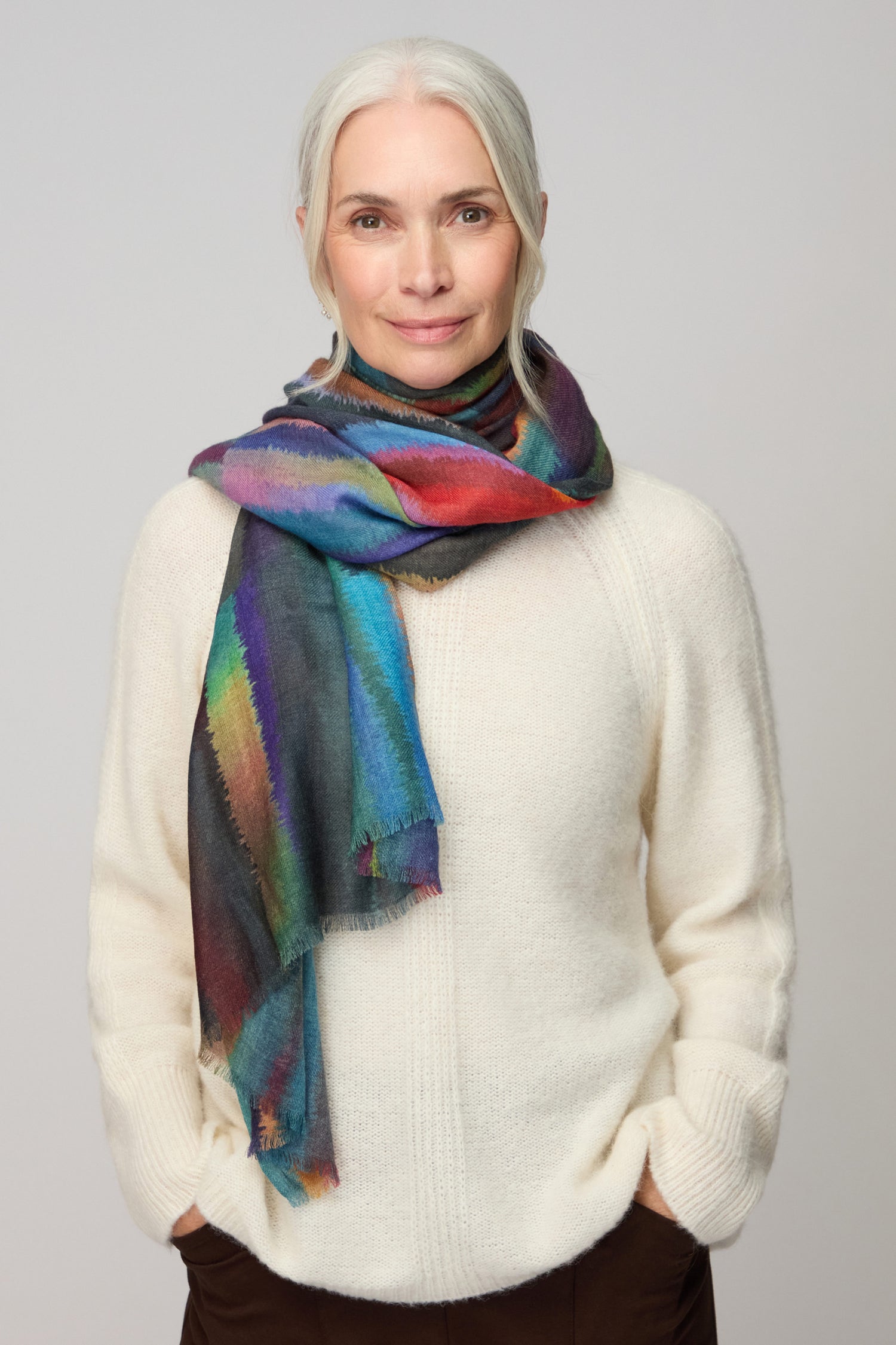 A woman with white hair stands against a plain light background, wearing a cream sweater and the Fluid Stripe Wool Scarf, with her hands in her pockets.