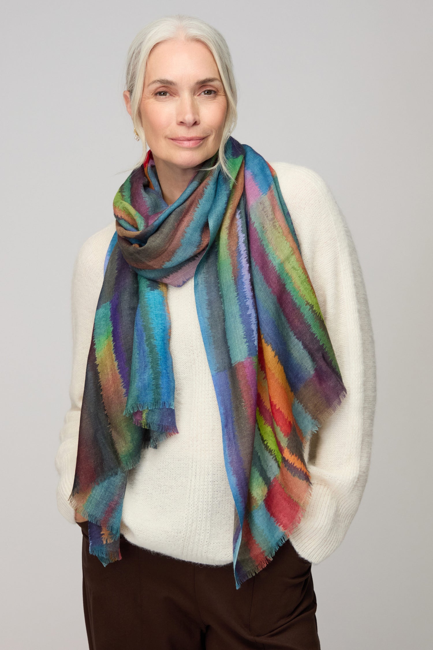 An older woman with gray hair, in a white sweater and brown pants, stands against a plain background wearing the Fluid Stripe Wool Scarf.