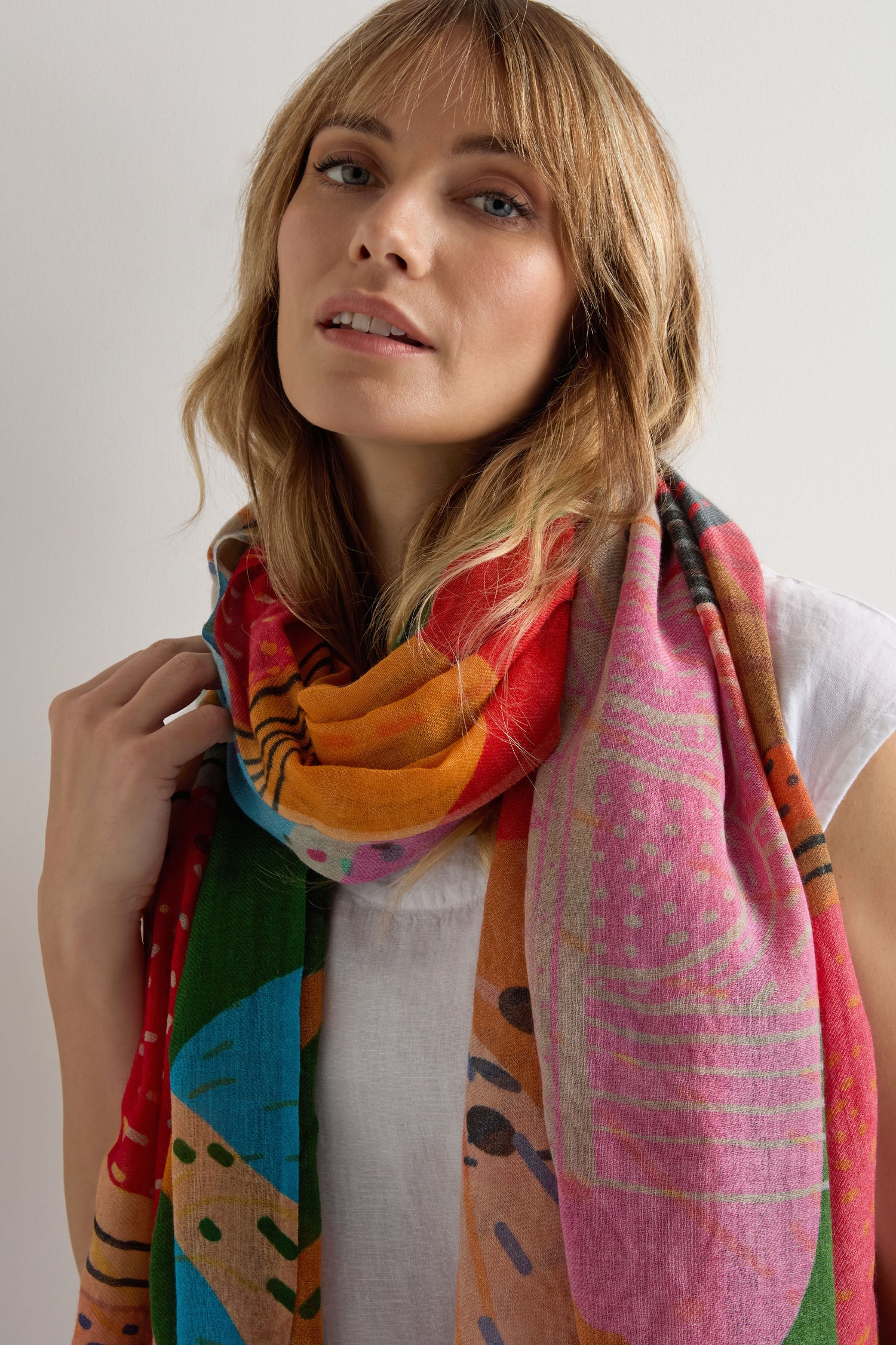 A woman wears a white top and the Tutti Frutti Wool Twill Scarf, featuring vibrant patterns in wool and silk, standing against a plain white background.