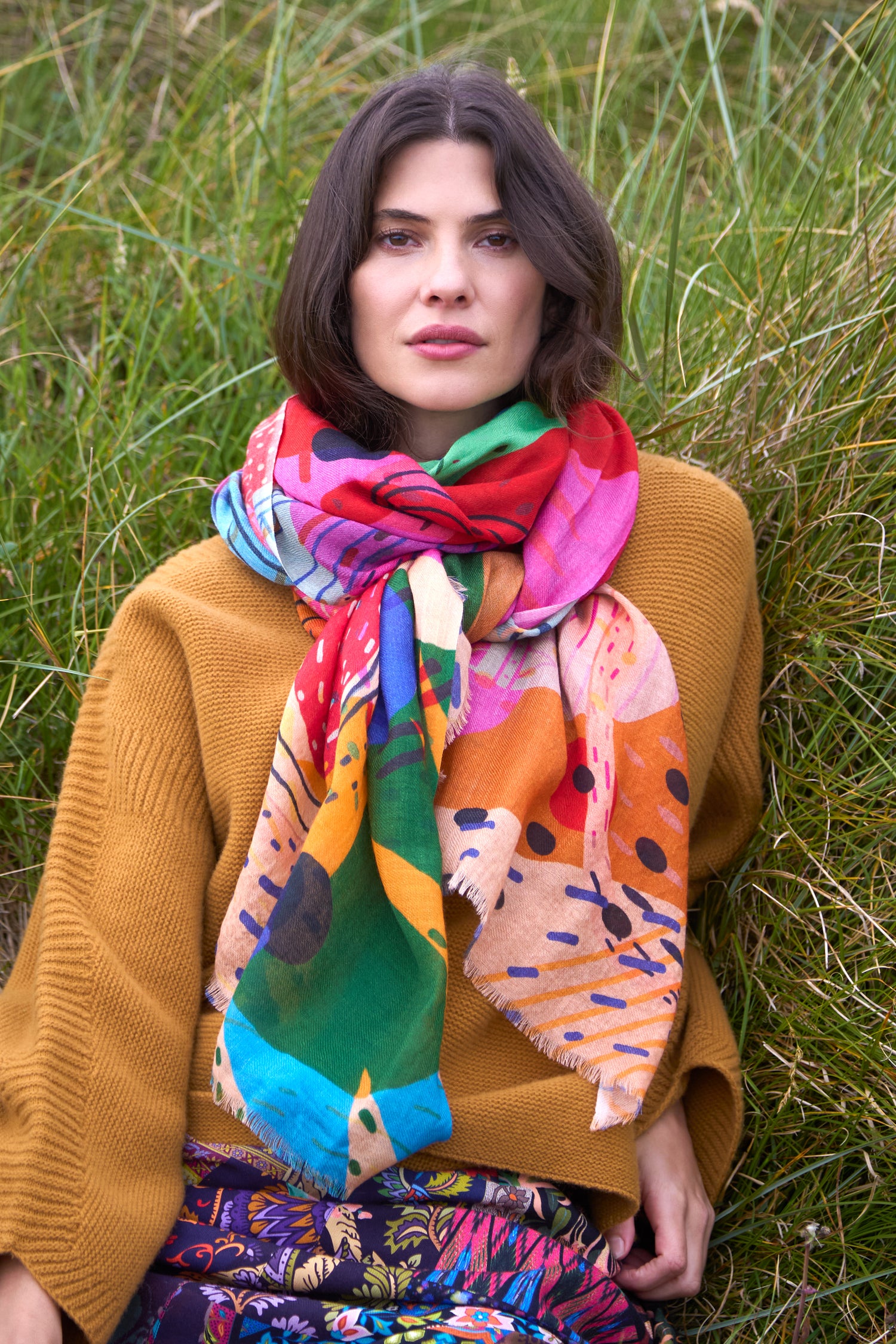 A woman with shoulder-length brown hair wears a mustard sweater and a floral skirt, sitting in tall grass, accessorized with the Tutti Frutti Wool Twill Scarf.