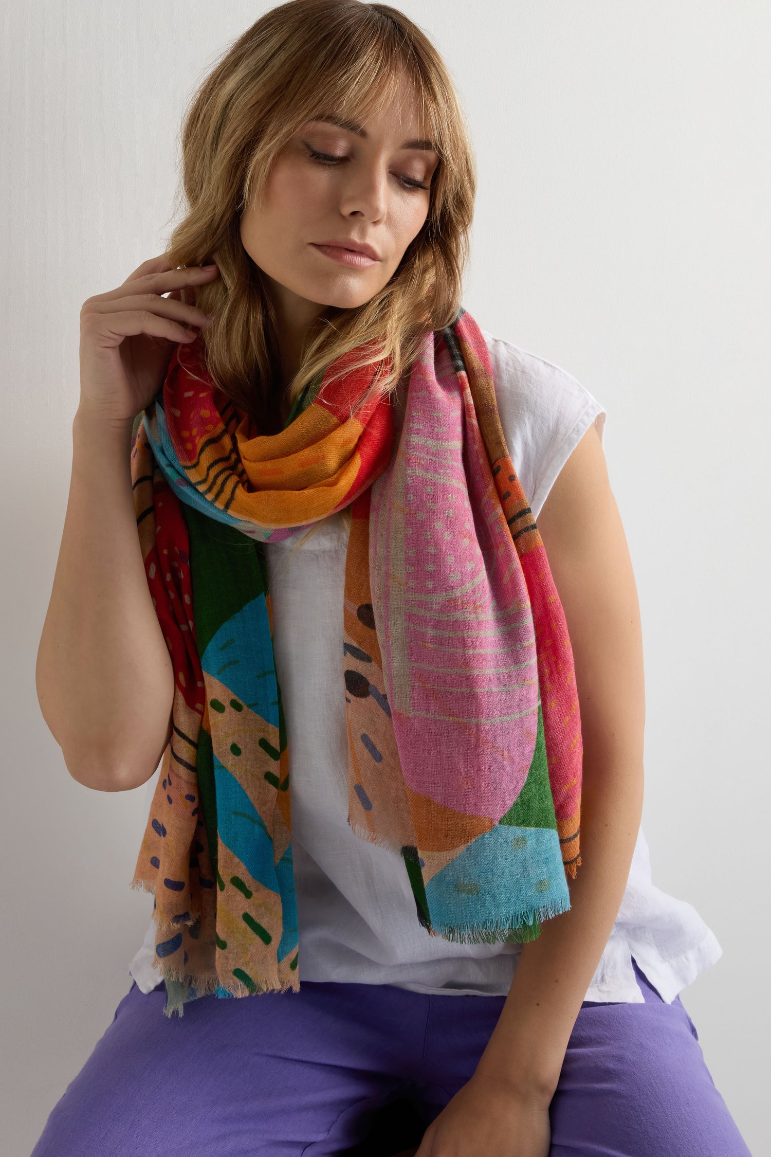 A woman with light brown hair wears a white short-sleeve top, purple pants, and the Tutti Frutti Wool Twill Scarf with a vibrant abstract design in wool and silk, seated against a plain light background.