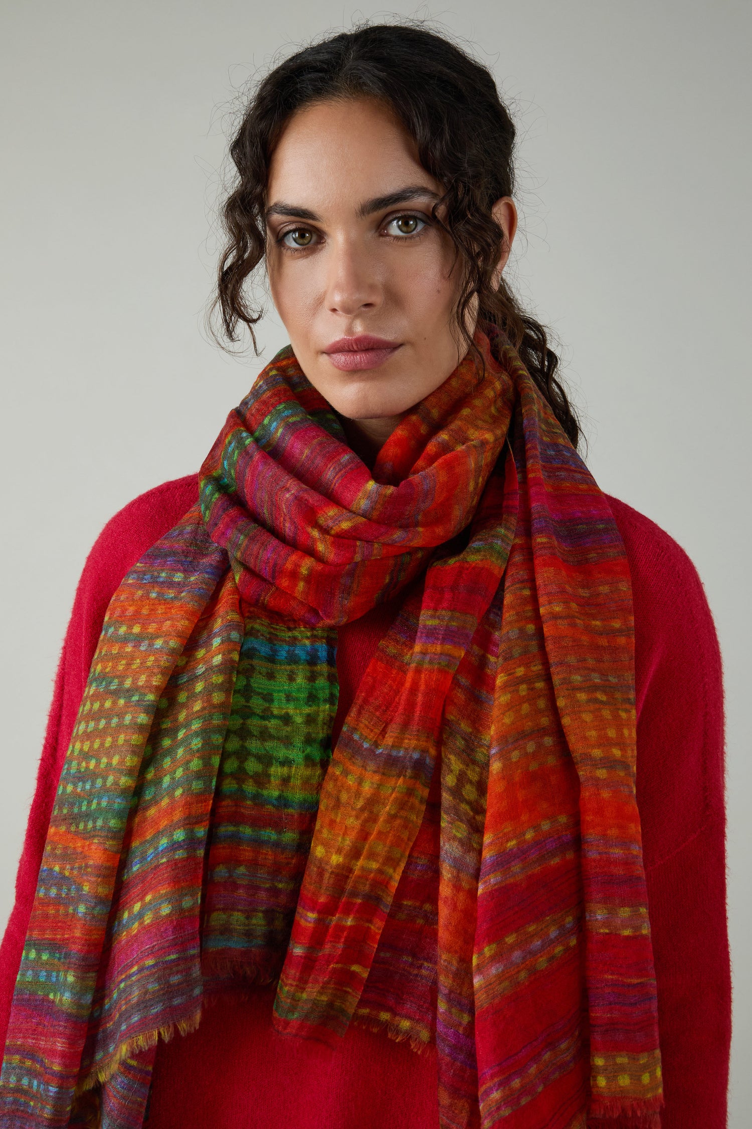 Woman with wavy brown hair wears a red sweater and the Sundown Stripe Wool Scarf, standing against a plain light gray background.