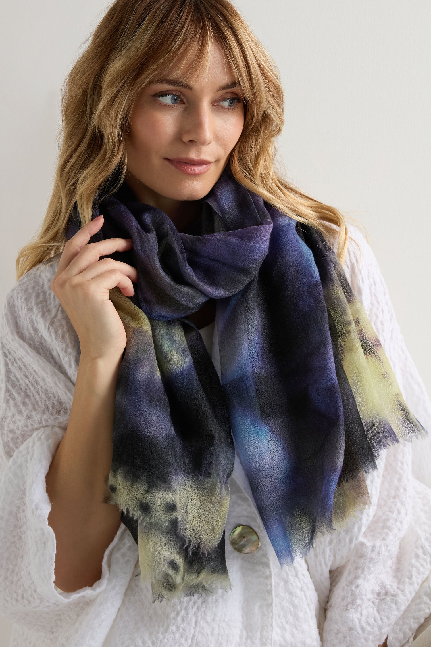 A woman stands against a plain light background wearing a textured white top and the Spiral Tie Dye Wool Scarf in blue, purple, and yellow.