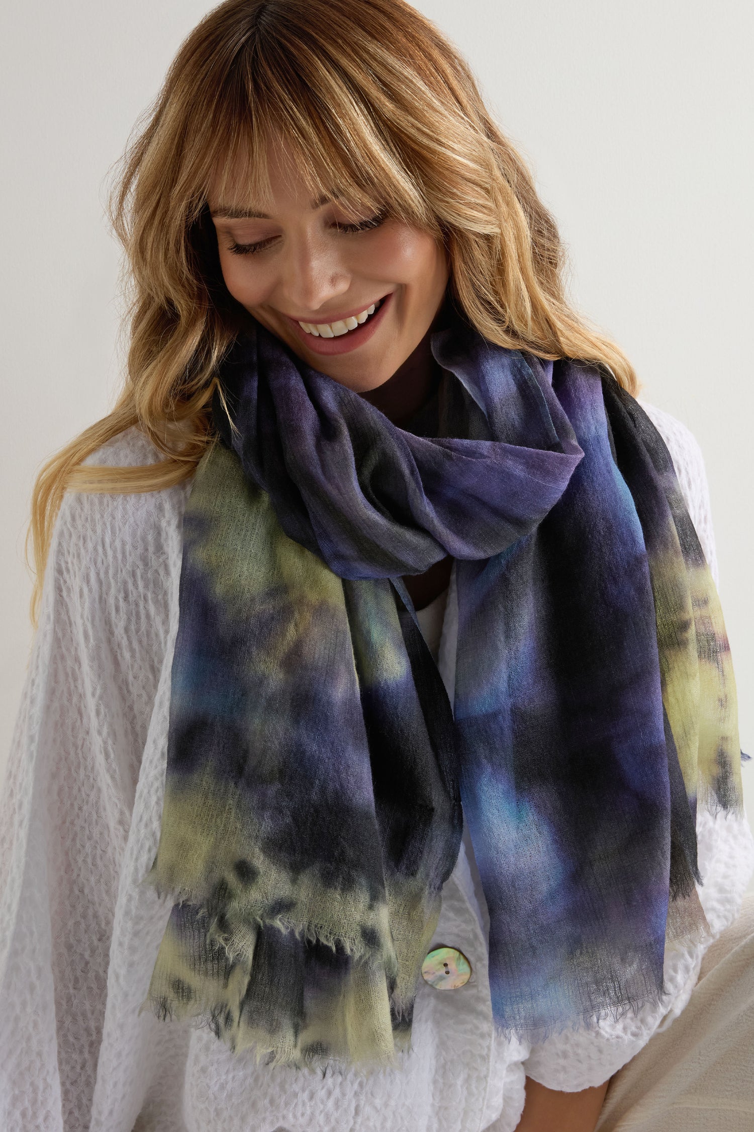 A woman smiles while looking down, wearing a white textured top and the Spiral Tie Dye Wool Scarf in vibrant blue, black, green, and yellow tones against a plain background.