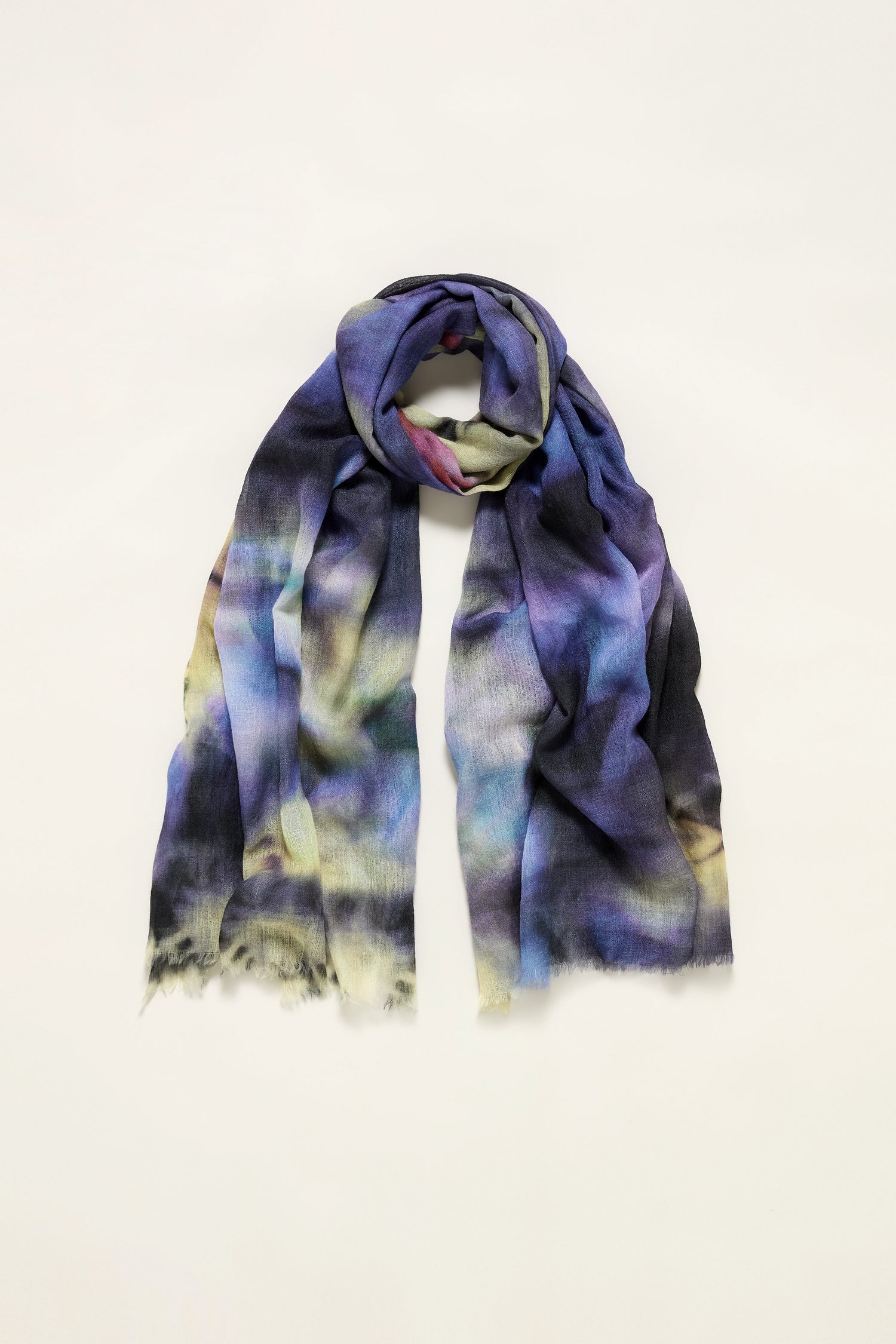 The Spiral Tie Dye Wool Scarf features a blend of blue, purple, yellow, and black tones in a multicolored spiral design, shown loosely folded and laid flat on a plain light background.