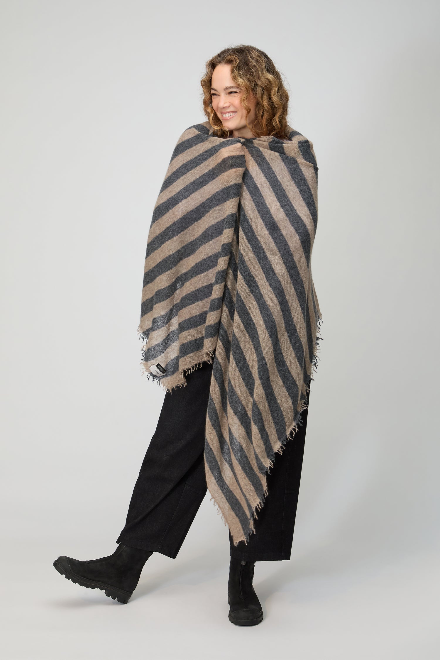A woman smiles, wrapped in the Soft Stripe Cashmere Scarf with fringed edges. She pairs it with black pants and boots, creating a cozy winter look against a plain background.
