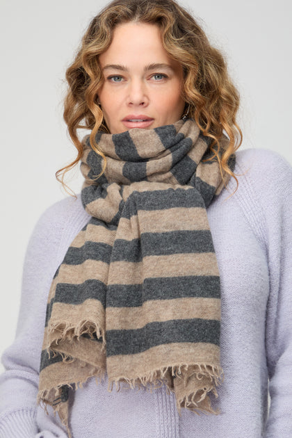 A woman with wavy hair wears a light purple sweater and the Soft Stripe Cashmere Scarf in beige and gray, featuring fringed edges—an elegant accessory to elevate any winter wardrobe.