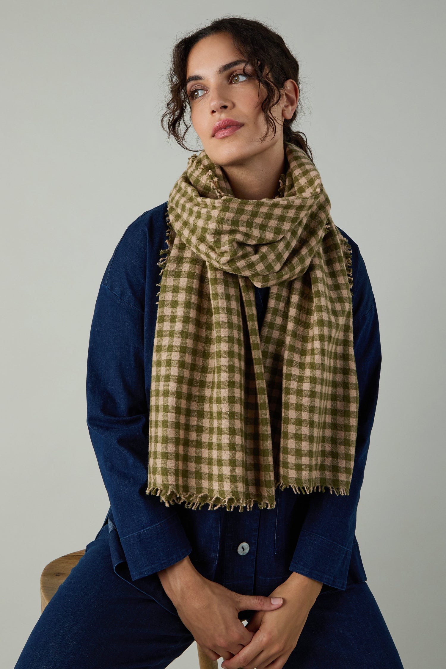 A woman in blue denim sits on a stool against a plain background, wearing the Boiled Wool Gingham Check Scarf in green and beige for lightweight warmth.