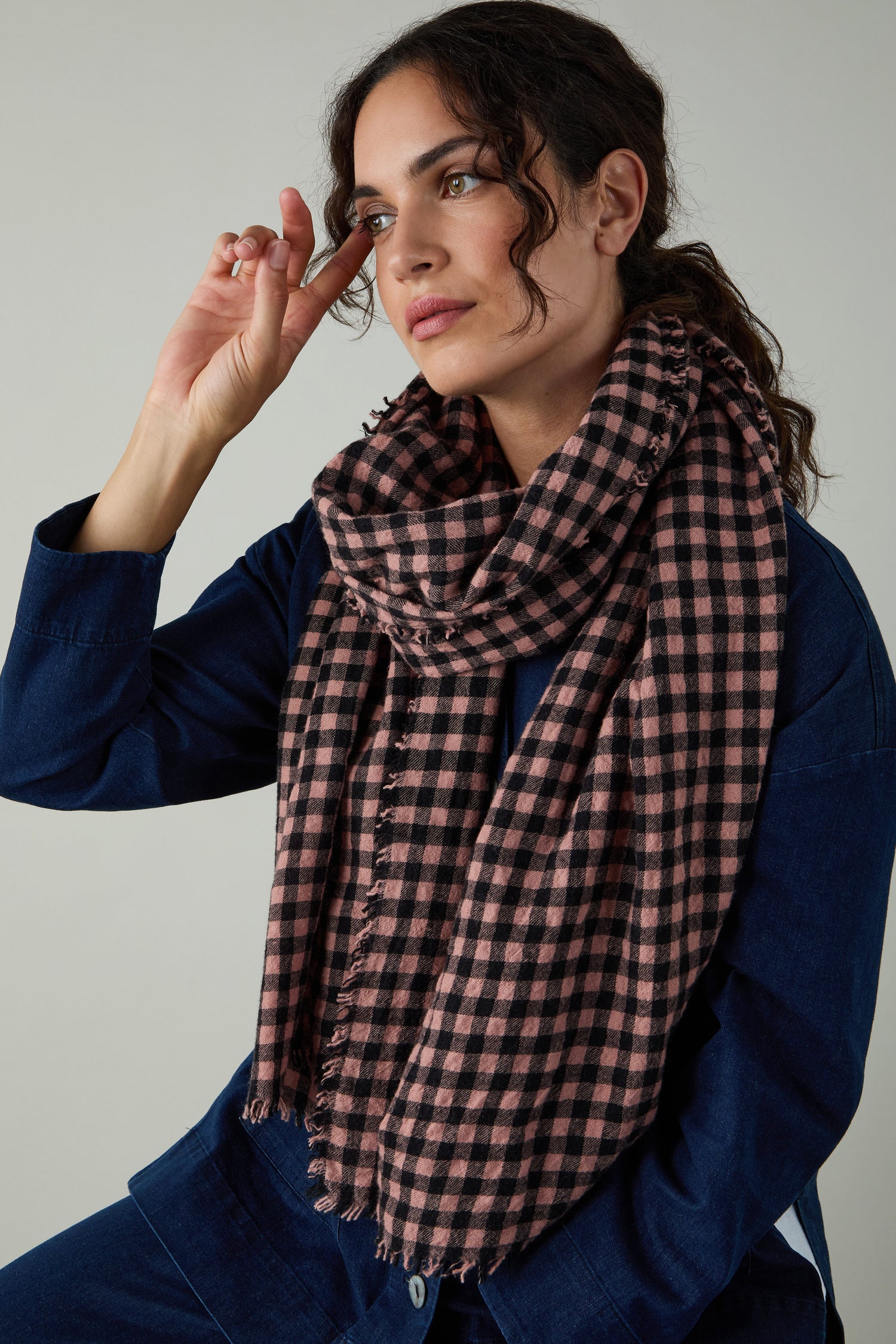 A woman in blue sits with one hand near her face, wearing the Boiled Wool Gingham Check Scarf—an effortless style statement that offers lightweight warmth.