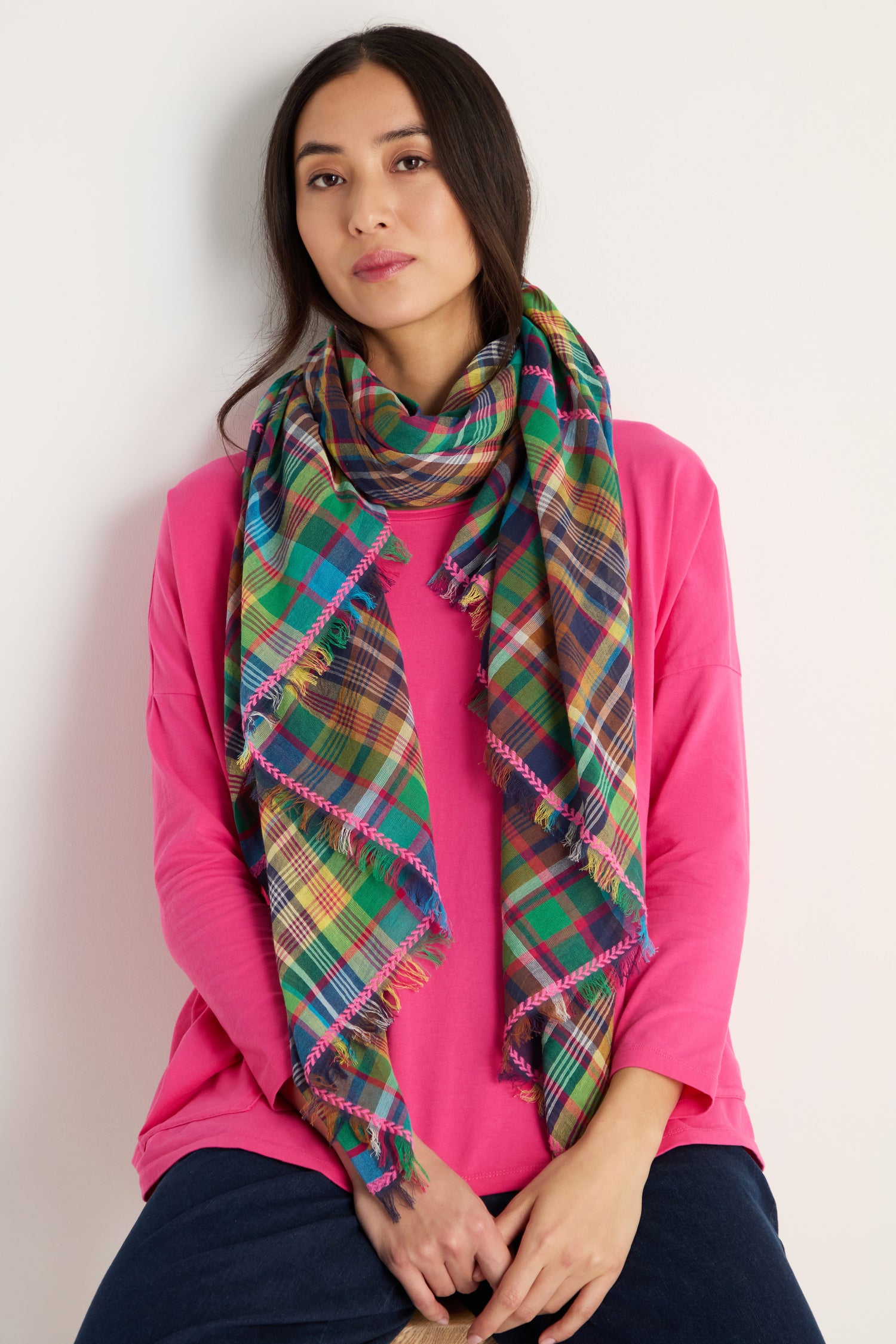 A woman in a bright pink top and dark pants wears the Fuchsia Embroidered Check Scarf draped around her neck, sitting against a plain light background.