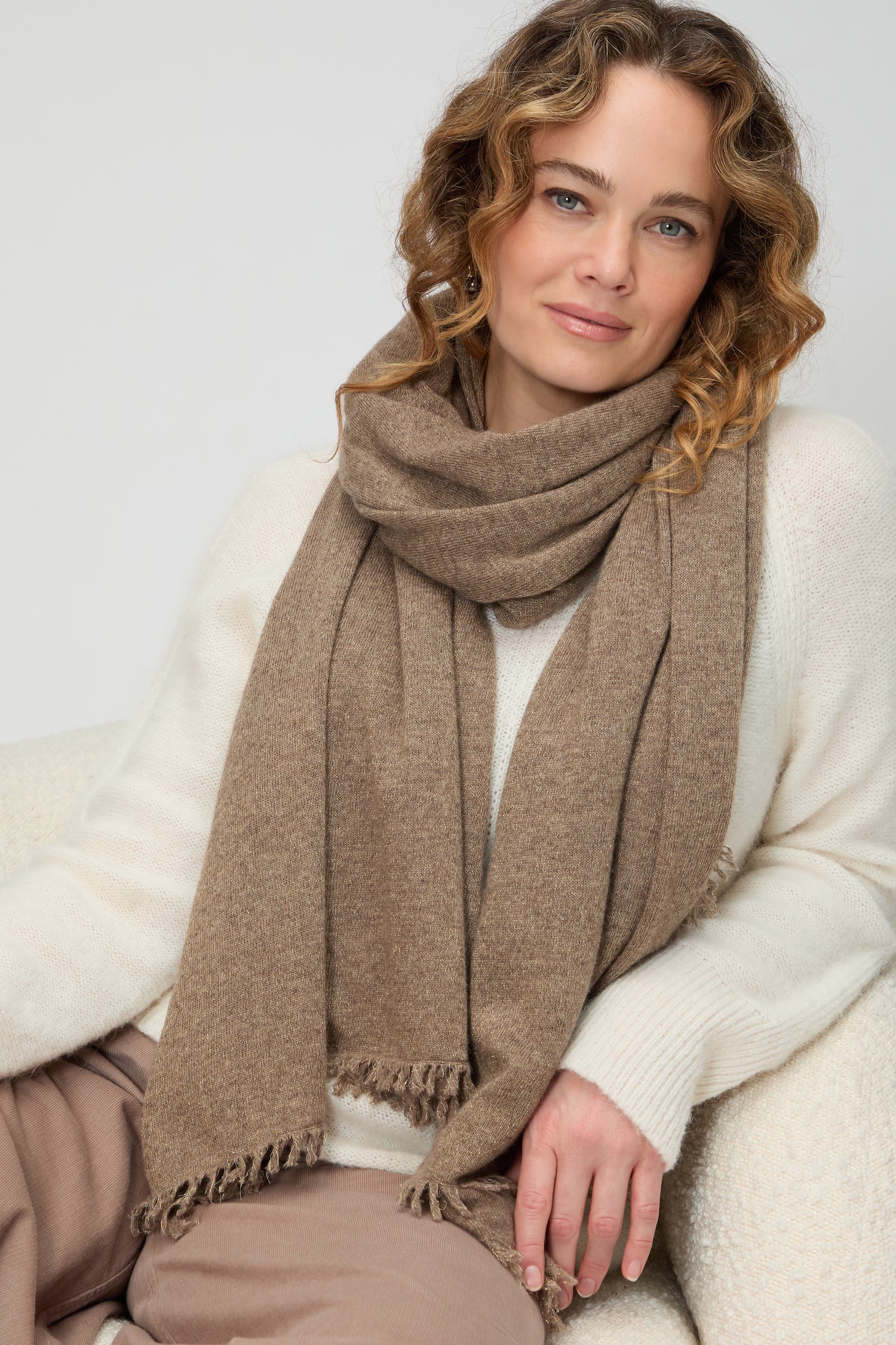 A woman with curly hair wears the Cashmere Sparkle Scarf and a cream sweater, seated on a light-colored chair against a plain background.