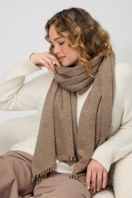 A woman with wavy hair and closed eyes wears a white sweater, brown pants, and the Cashmere Sparkle Scarf in neutral tones.