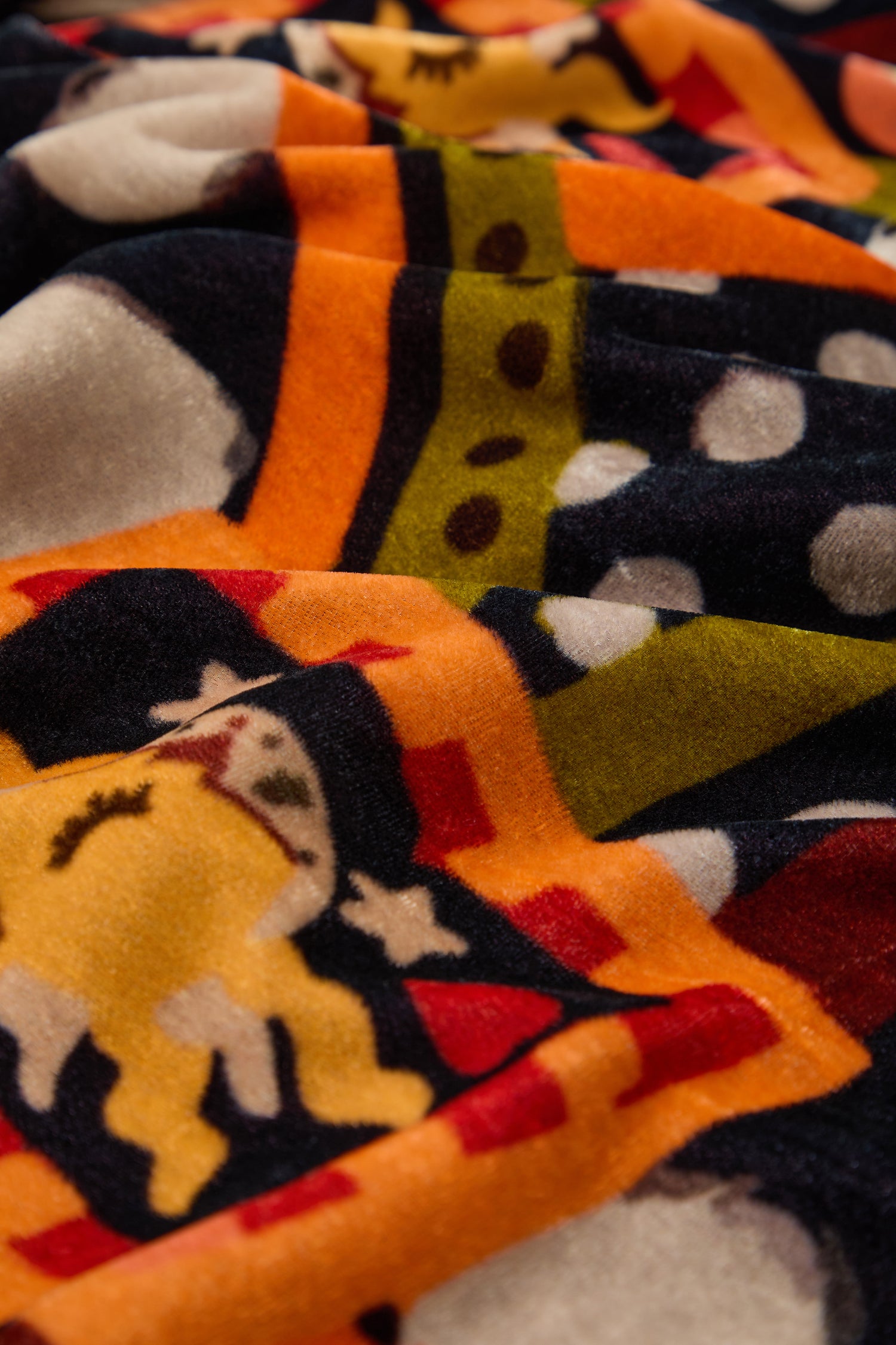 A close-up of the Hera Velvet Scarf showcases colorful cartoon patterns in yellow, orange, green, black, and white, giving it a playful charm reminiscent of jewel tones and stylish eveningwear accessories.