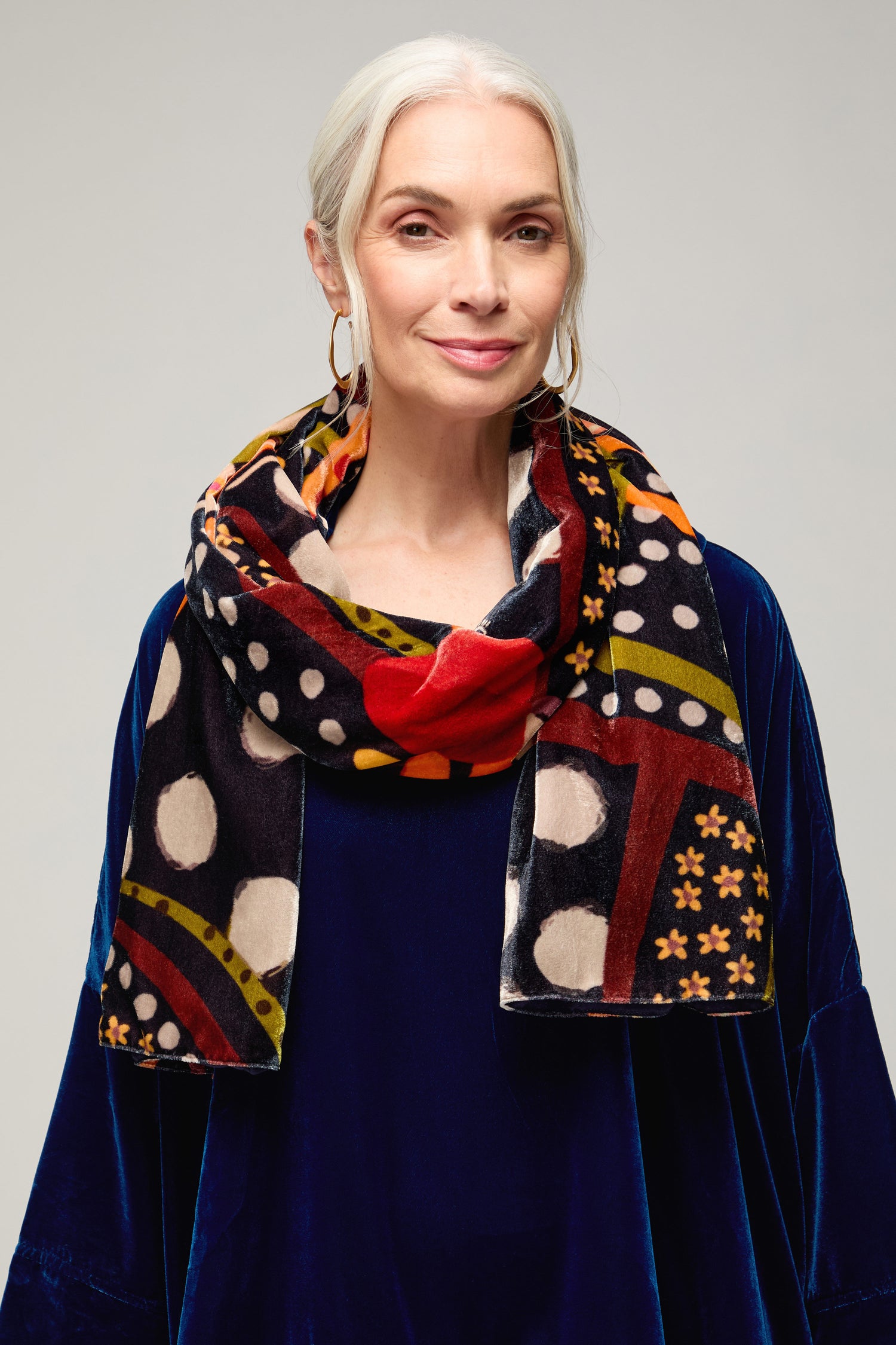 An older woman with silver hair and hoop earrings wears the Hera Velvet Scarf, which features traditional motifs, paired with a velvet navy top against a neutral background.