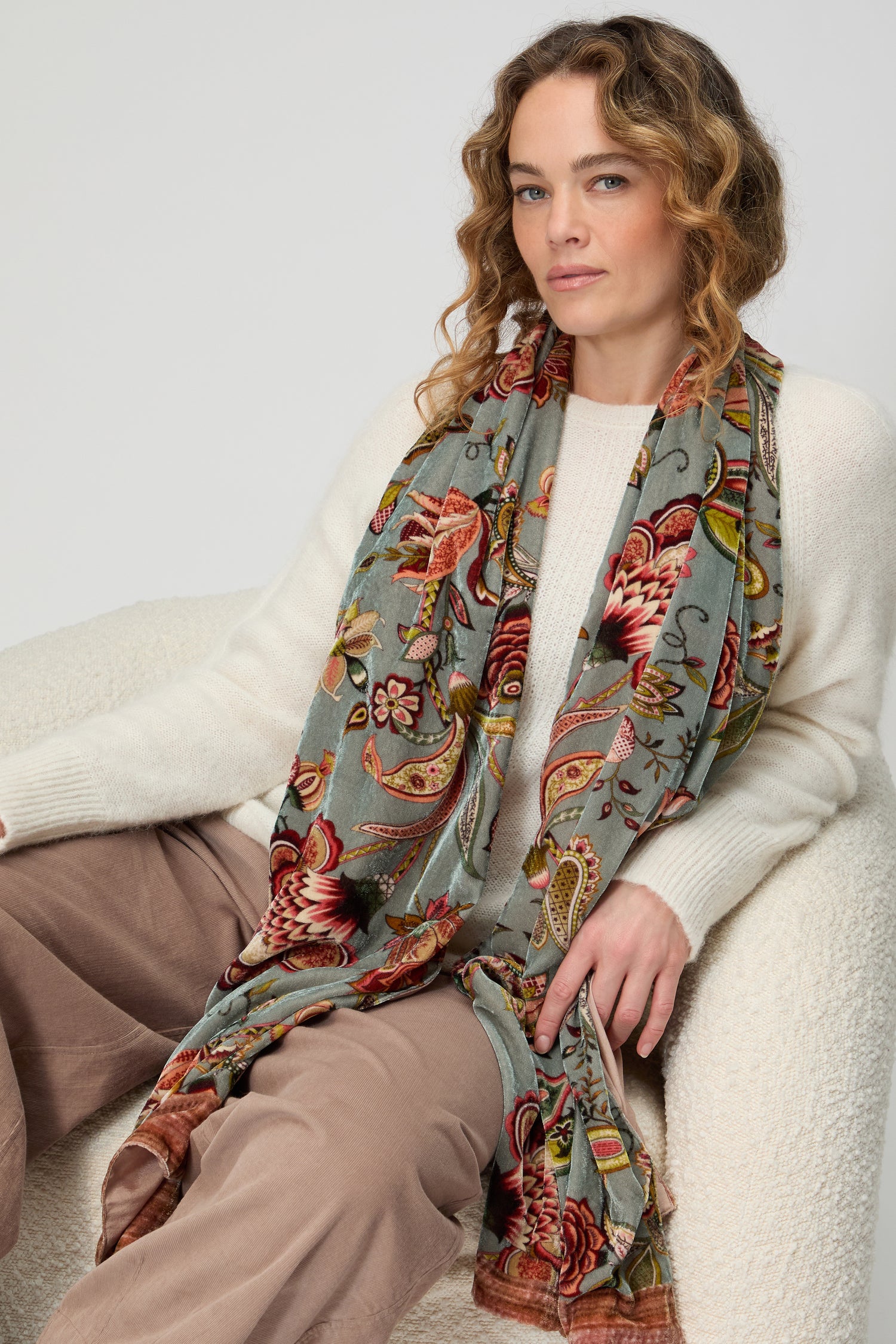 A woman with wavy hair wears a cream sweater, taupe pants, and the In Bloom Velvet Scarf as a statement accessory while sitting on a textured white chair against a neutral background.