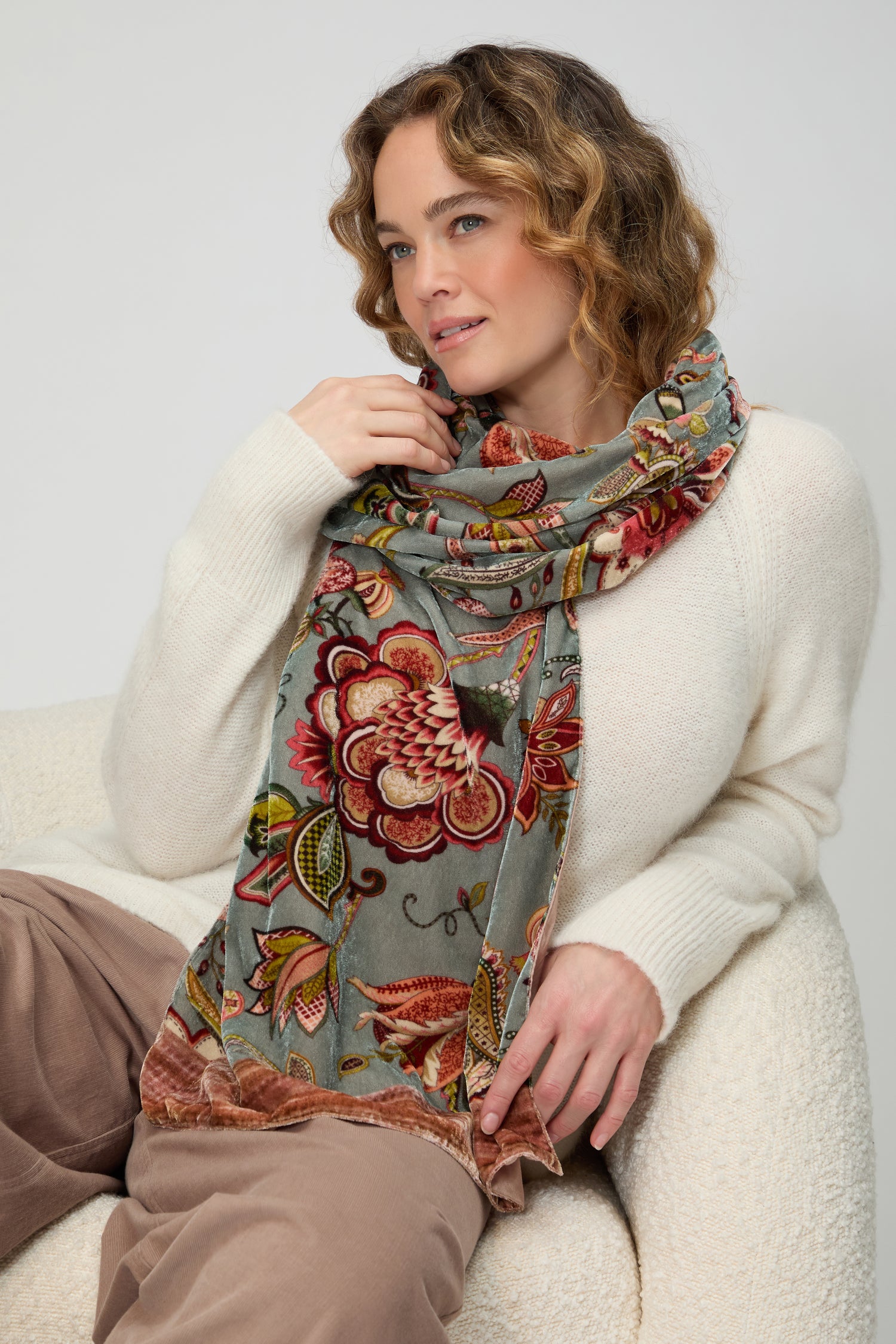 A woman seated on a light chair wears a cream sweater, taupe pants, and the In Bloom Velvet Scarf wrapped around her neck as a subtle statement accessory, looking to the side.