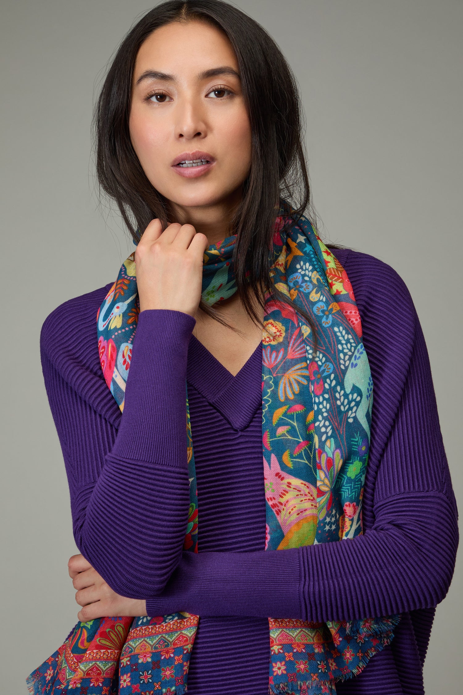 A woman with long dark hair, wearing a purple sweater and the Enchanted Woodland Wool Mix Scarf with botanical motifs, poses with one hand near her face against a plain gray background.