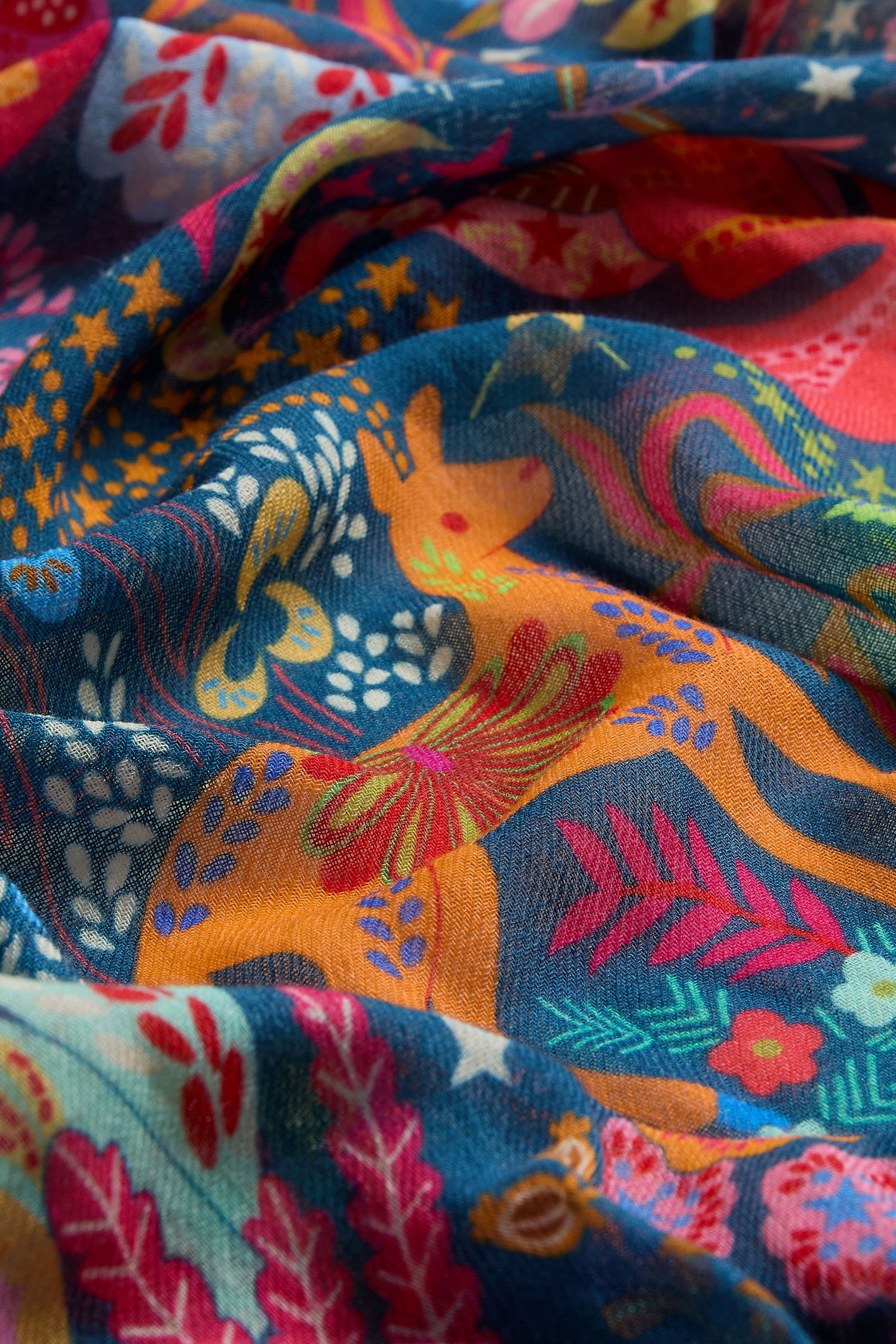The Enchanted Woodland Wool Mix Scarf features a vibrant folkloric pattern of deer, leaves, flowers, and stars in orange, red, blue, and yellow—with botanical accents—perfect as a lightweight accessory.