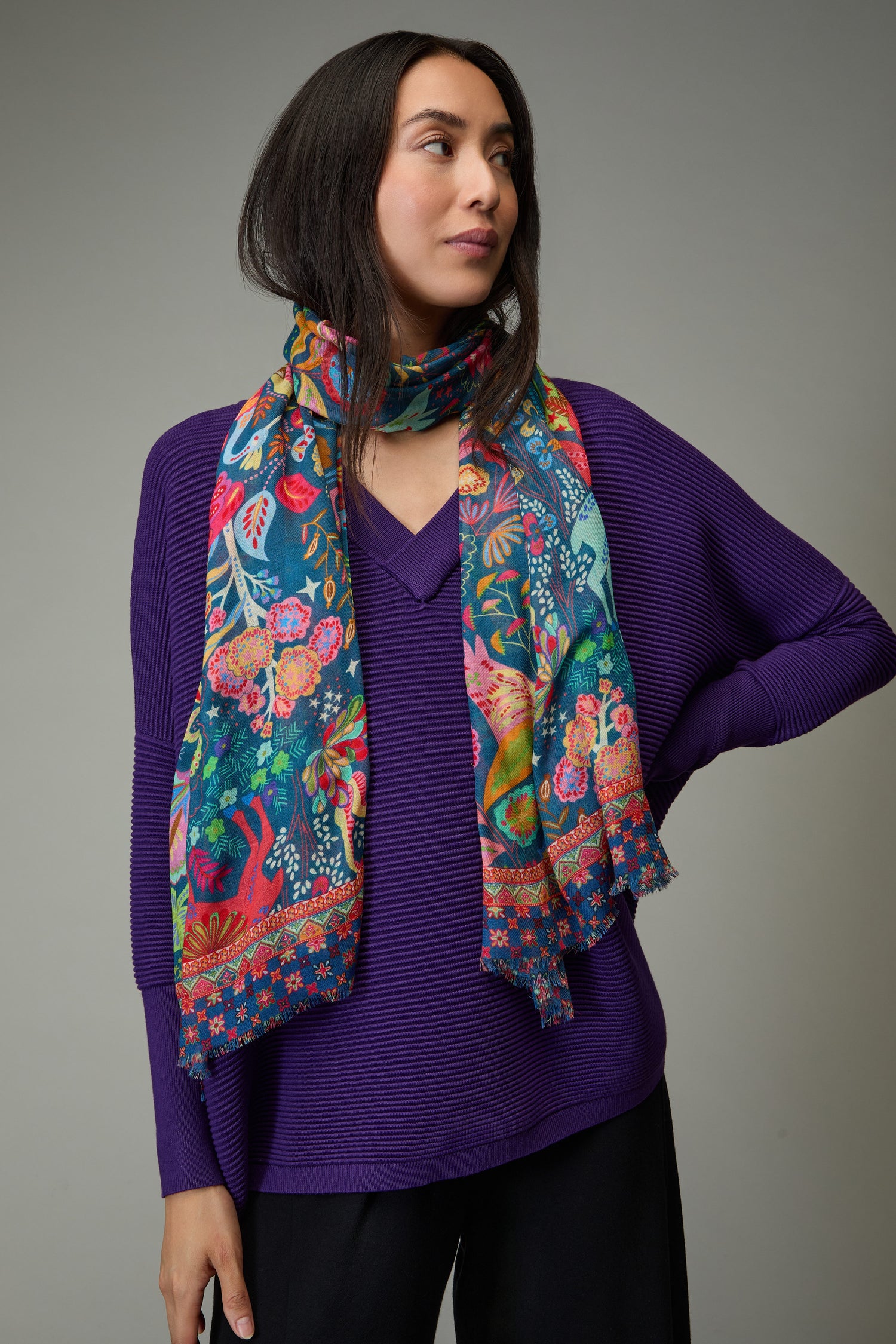 A woman with long dark hair wears a purple sweater and the Enchanted Woodland Wool Mix Scarf, featuring colorful botanical motifs, standing against a plain gray background.