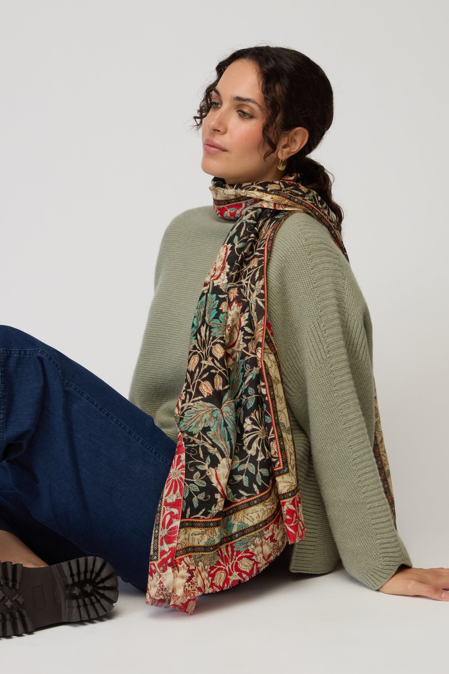 A woman in a green sweater, blue jeans, and black boots sits on the floor with the Ornamental Floral Wool Mix Scarf draped over her shoulder against a plain background.
