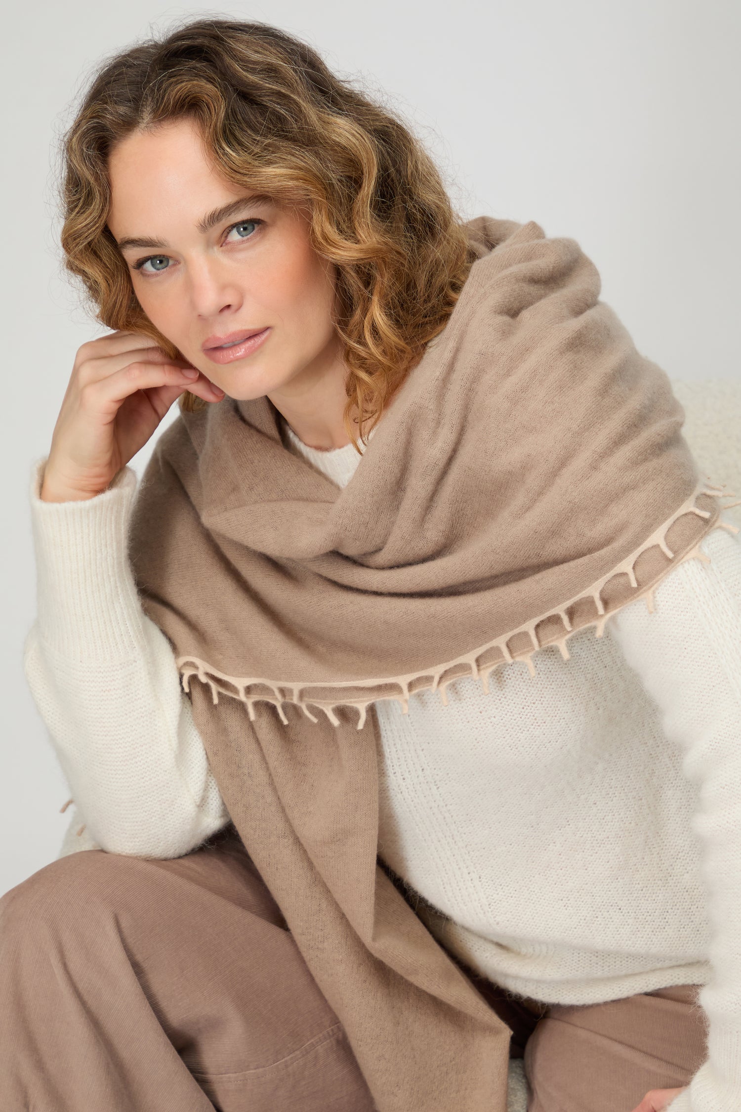 Woman with wavy hair wears a Contrast Trim Cream Cashmere Scarf over a white sweater and brown pants, sitting with her hand resting against her face.