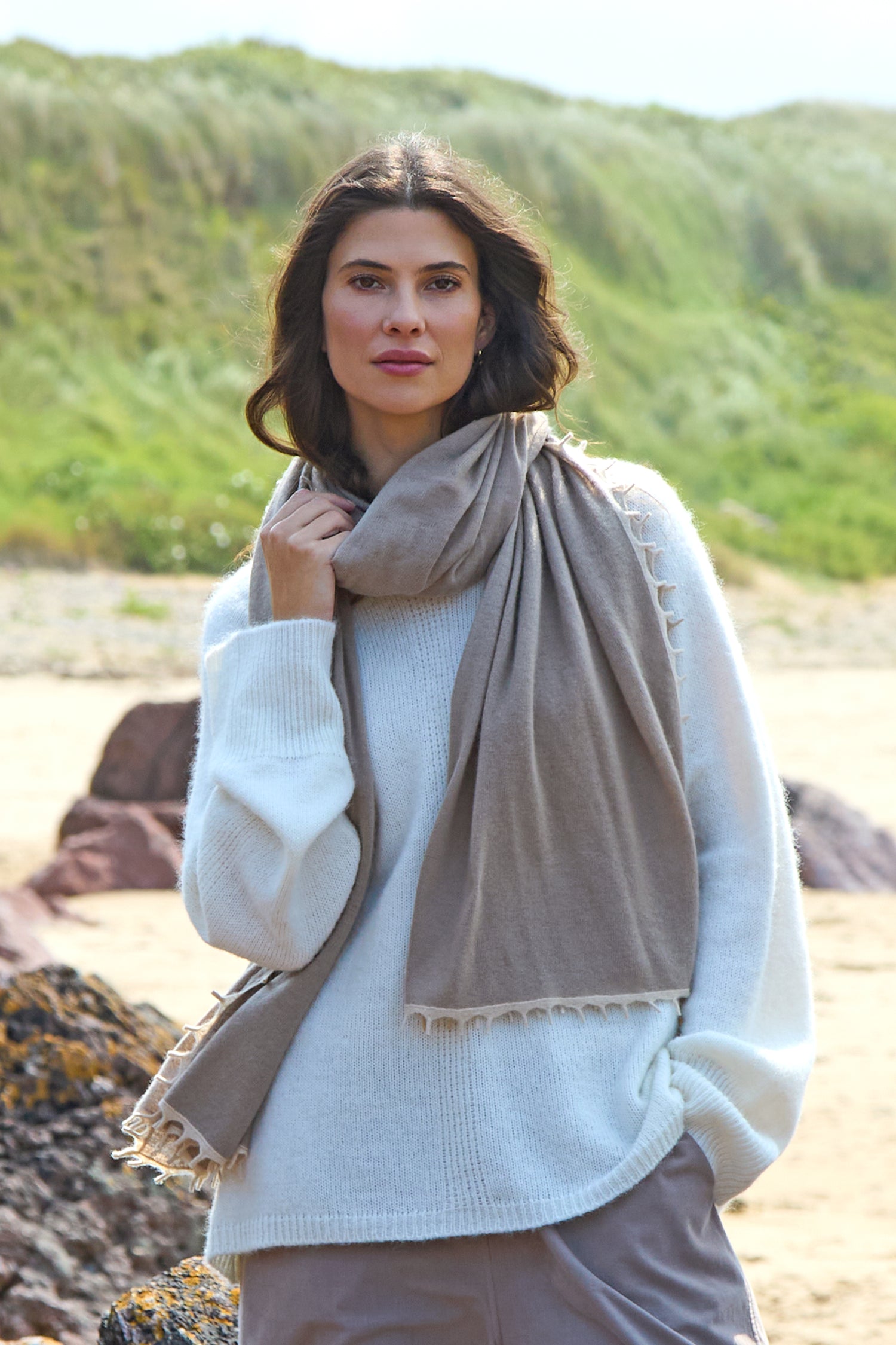 A woman enjoys the fresh sea breeze on a beach, wearing a white sweater and the Contrast Trim Cream Cashmere Scarf, with greenery and sand dunes in the background.
