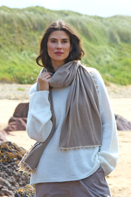 A woman enjoys the fresh sea breeze on a beach, wearing a white sweater and the Contrast Trim Cream Cashmere Scarf, with greenery and sand dunes in the background.