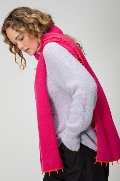 Wearing a light purple sweater and black pants, a person styles the Contrast Trim Pink Cashmere Scarf—bright pink with orange trim—while looking down with one hand in their pocket against a plain background.