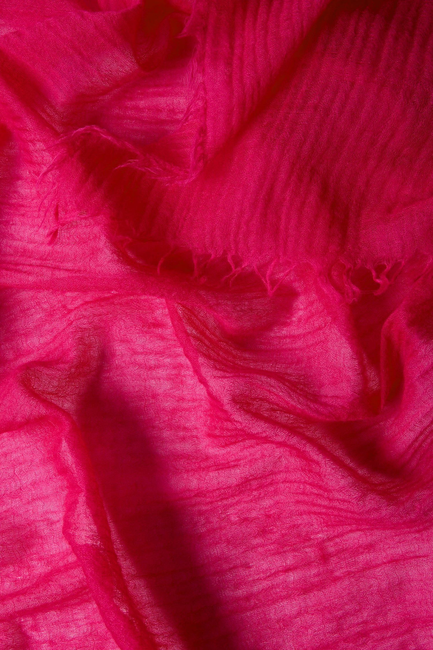 Close-up view of the Hibiscus Fine Wool Fringe Scarf, showcasing its bright pink hue, textured lightweight fabric, visible creases, and soft frayed edges.