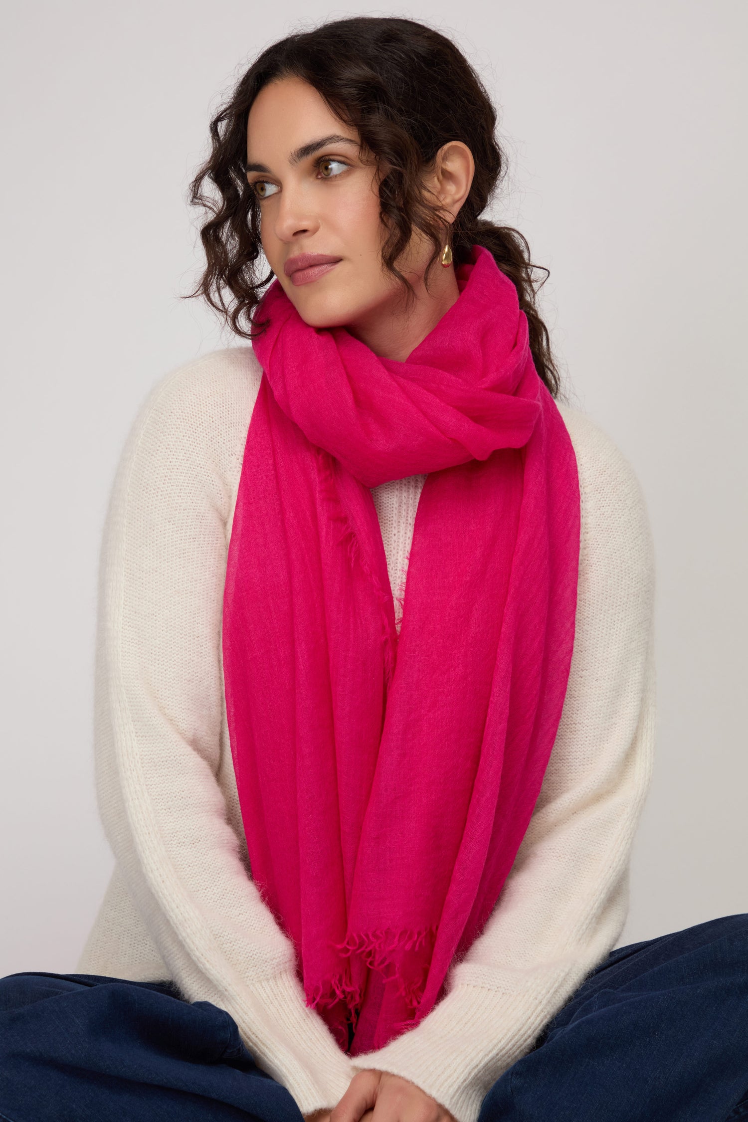 Woman with wavy dark hair wearing a white sweater and the Hibiscus Fine Wool Fringe Scarf, sitting with hands folded and looking slightly to the side against a plain background.