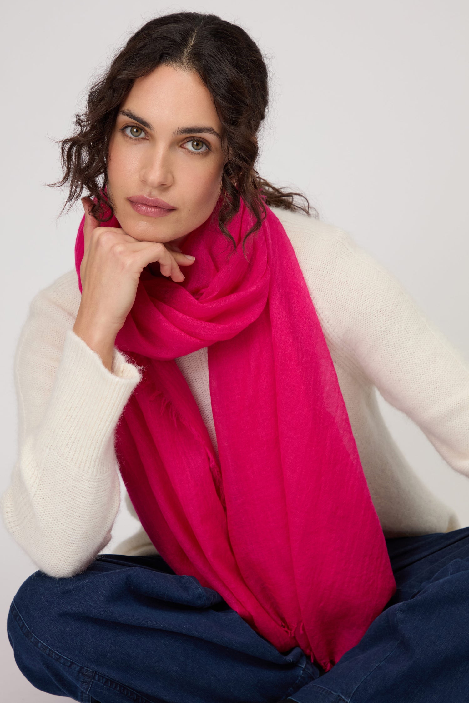 Woman with wavy dark hair in a white sweater and blue jeans, wearing the Hibiscus Fine Wool Fringe Scarf in fuchsia, sits with one hand resting against her face.