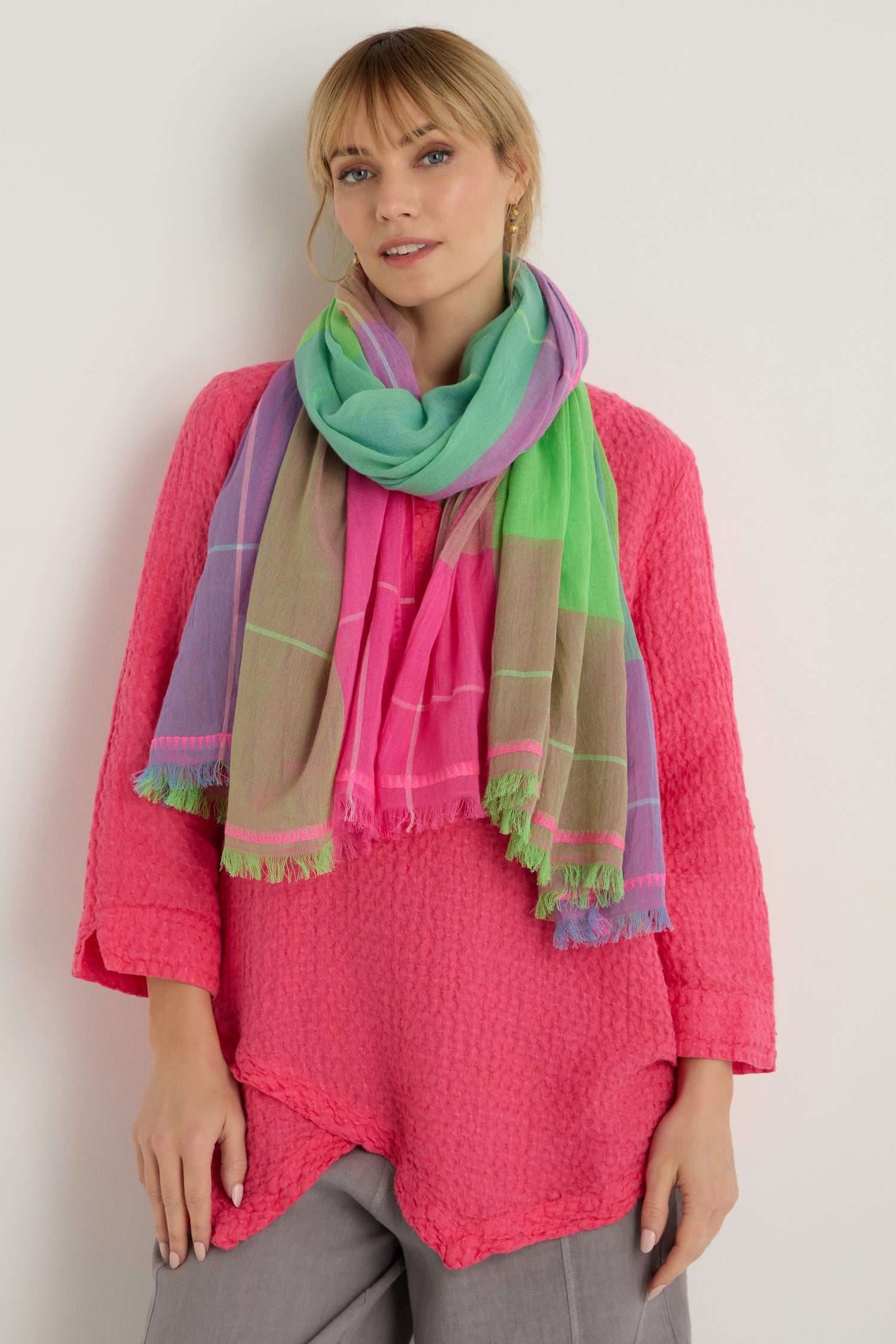 A woman stands against a white background wearing a bright pink textured top, gray pants, and the Embroidered Colour Pop Check Cotton Scarf with green, pink, and purple tones.