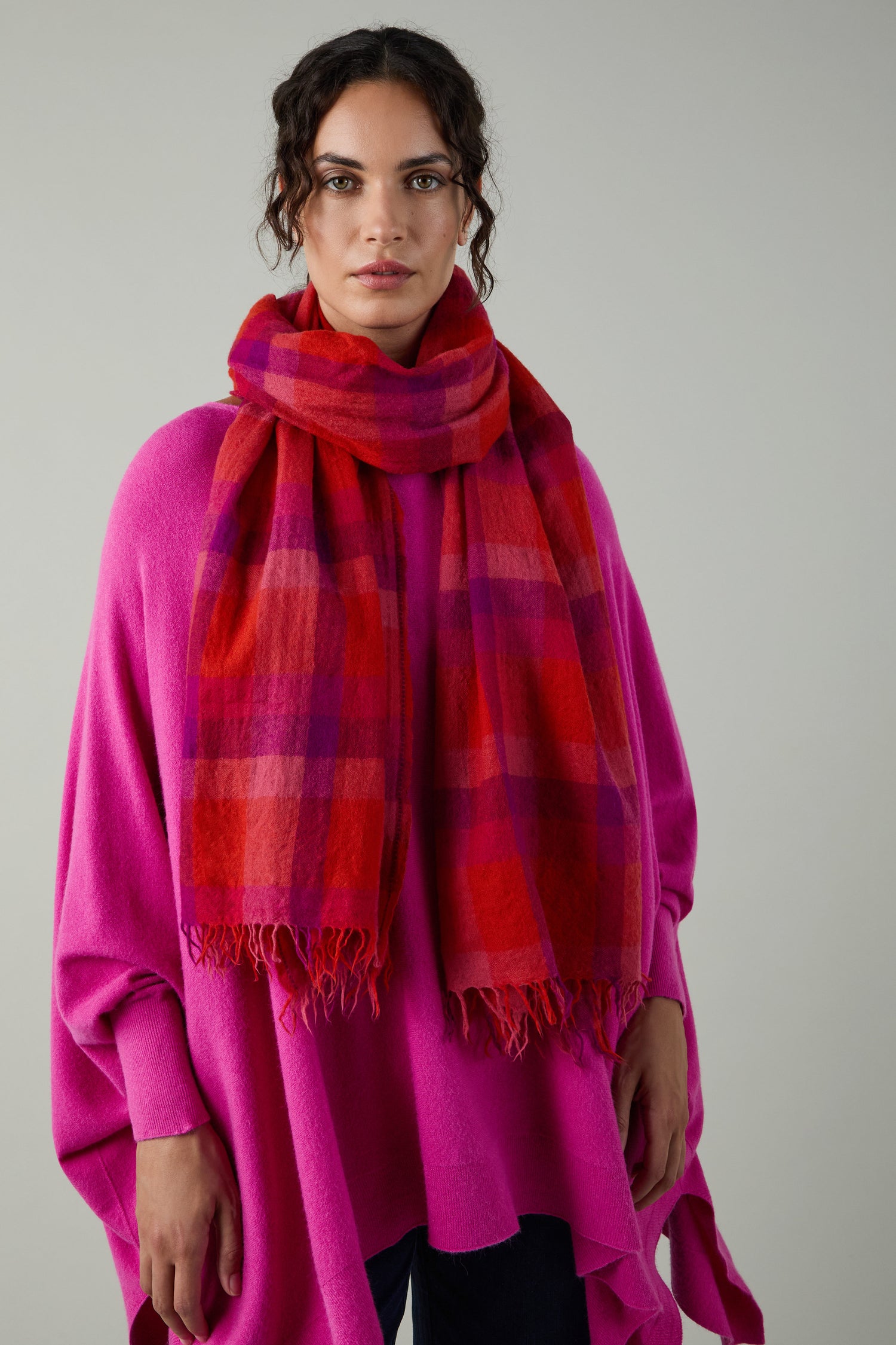 A person wears a bright pink poncho and the Sunset Check Wool Scarf, featuring bold red and pink plaid, standing against a plain light gray background.