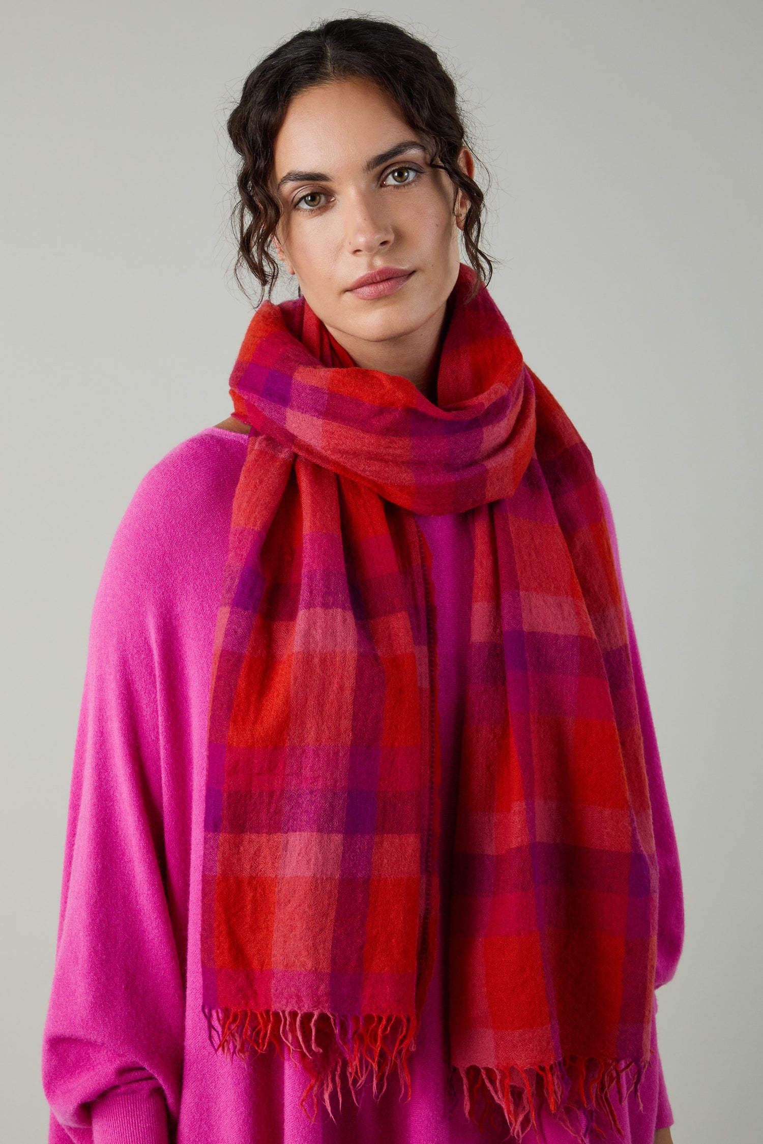 A woman wears the Sunset Check Wool Scarf, featuring red and pink plaid, with a bright pink top against a plain light gray background.