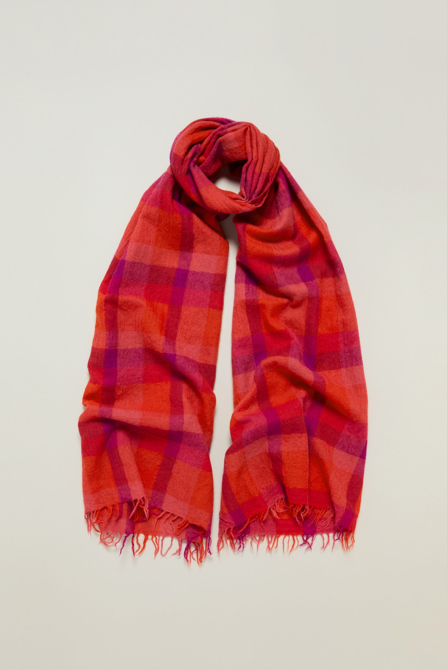 The Sunset Check Wool Scarf features red and pink checkered wool with frayed edges and subtle jewel tones, displayed flat on a light background.